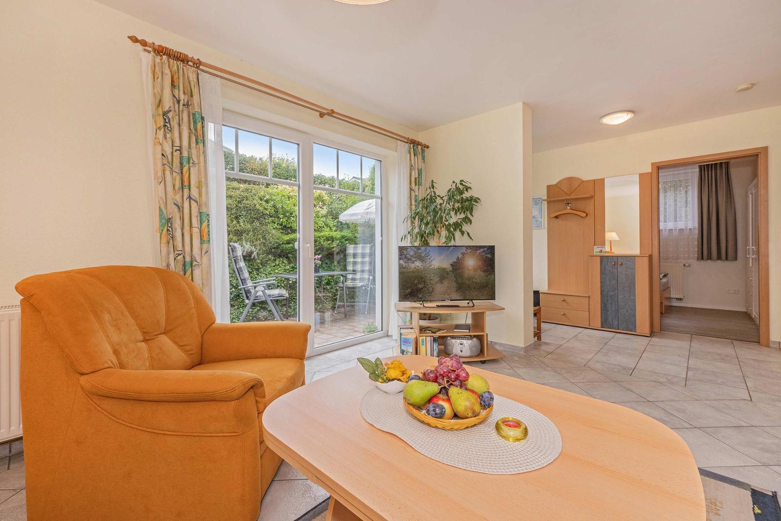 Wohnzimmer mit orangefarbenem Sessel, TV und Blick auf Balkon mit Garten.