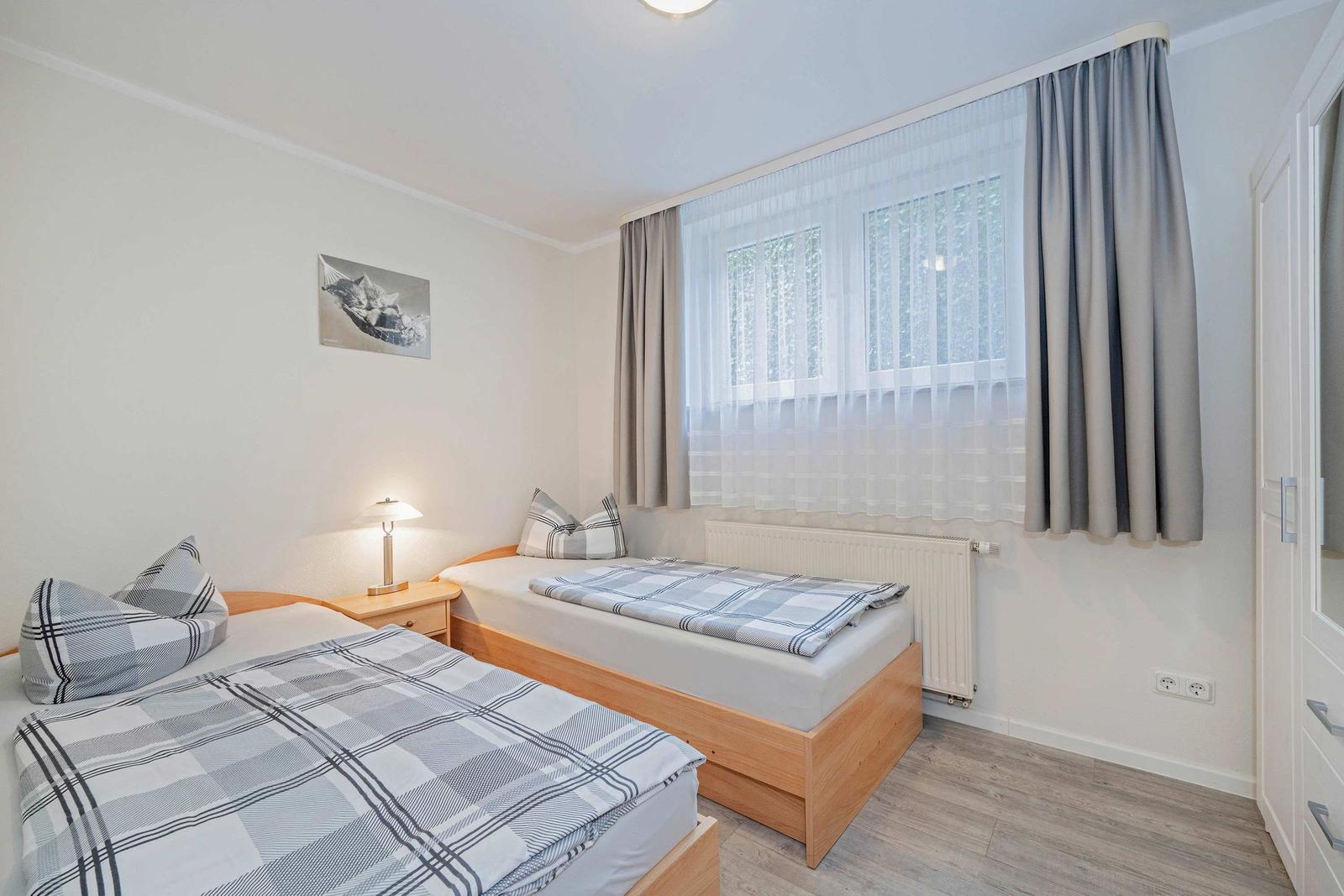 Zwei Einzelbetten mit karierten Bettwäsche, Fenster mit Vorhängen und Nachttisch mit Lampe.