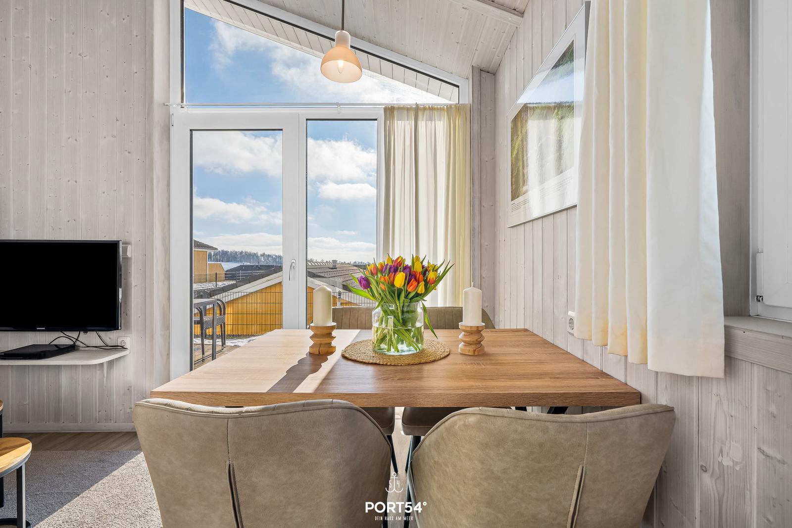 Dining area with wooden table, chairs, and flowers. Large windows with view of outdoor area.
