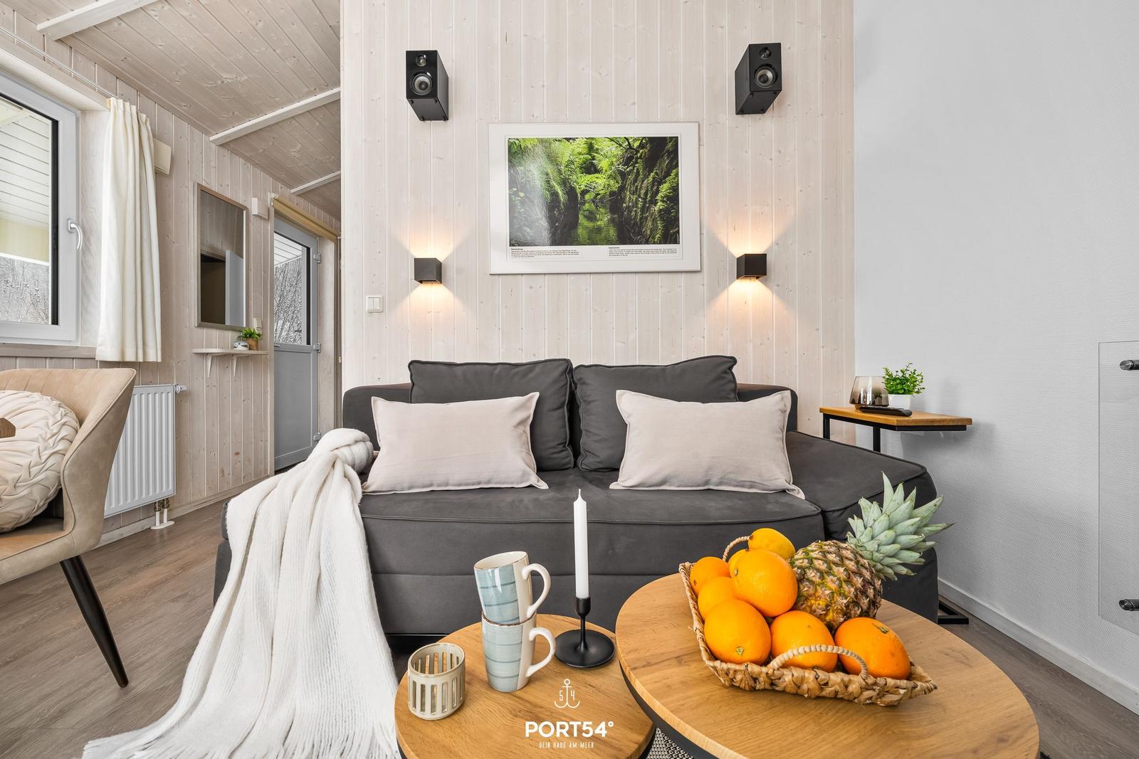 Ein Wohnzimmer mit grauem Sofa, Holztisch und Obst. Wandbild und Lautsprecher sind sichtbar.