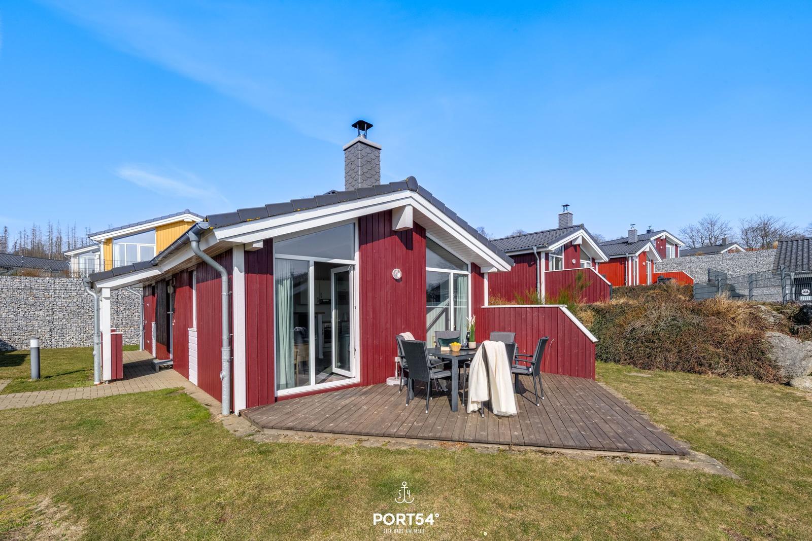 Rotes Holzhaus mit Terrasse und Sitzgelegenheit. Hinterhof mit Gras und Steinmauer.