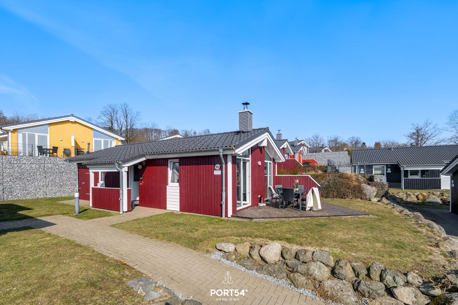 Rotes Ferienhaus mit Terrasse und Garten. Sicht auf Nachbarhäuser und klaren Himmel.