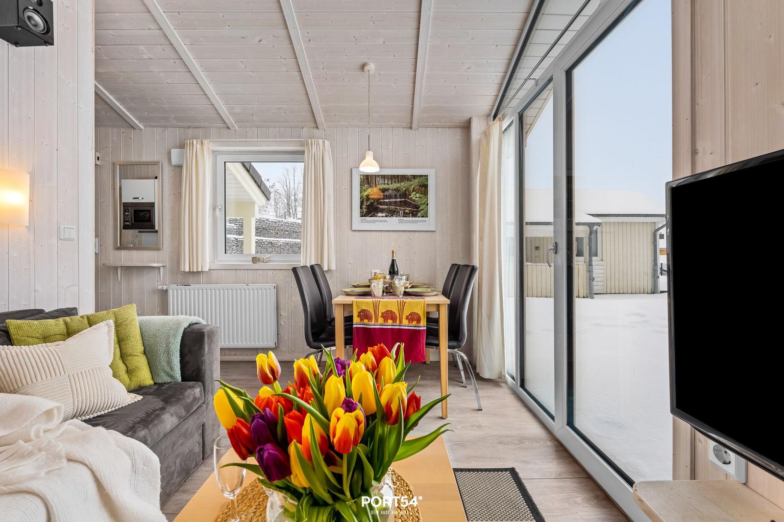 Wohnbereich mit Esstisch, Sofa und großen Fenstern. Blumen und TV sind sichtbar.