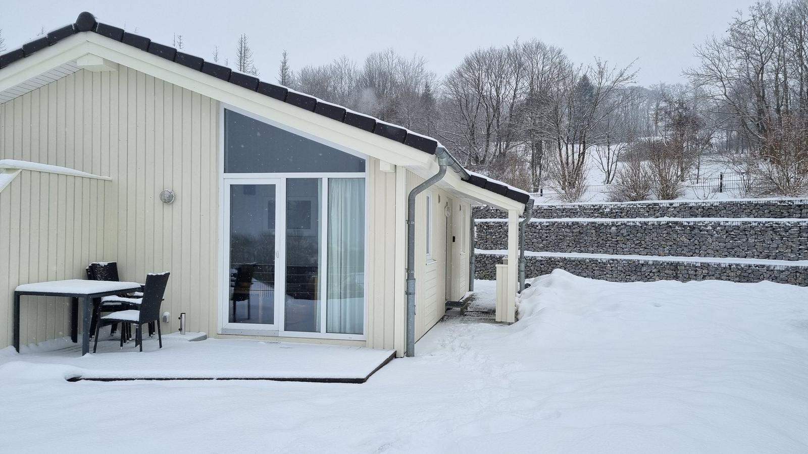 Wohnhaus mit Schneebedeckter Terrasse und Glasfront. Hintergrund: schneebedeckte Bäume und Steinmauer.