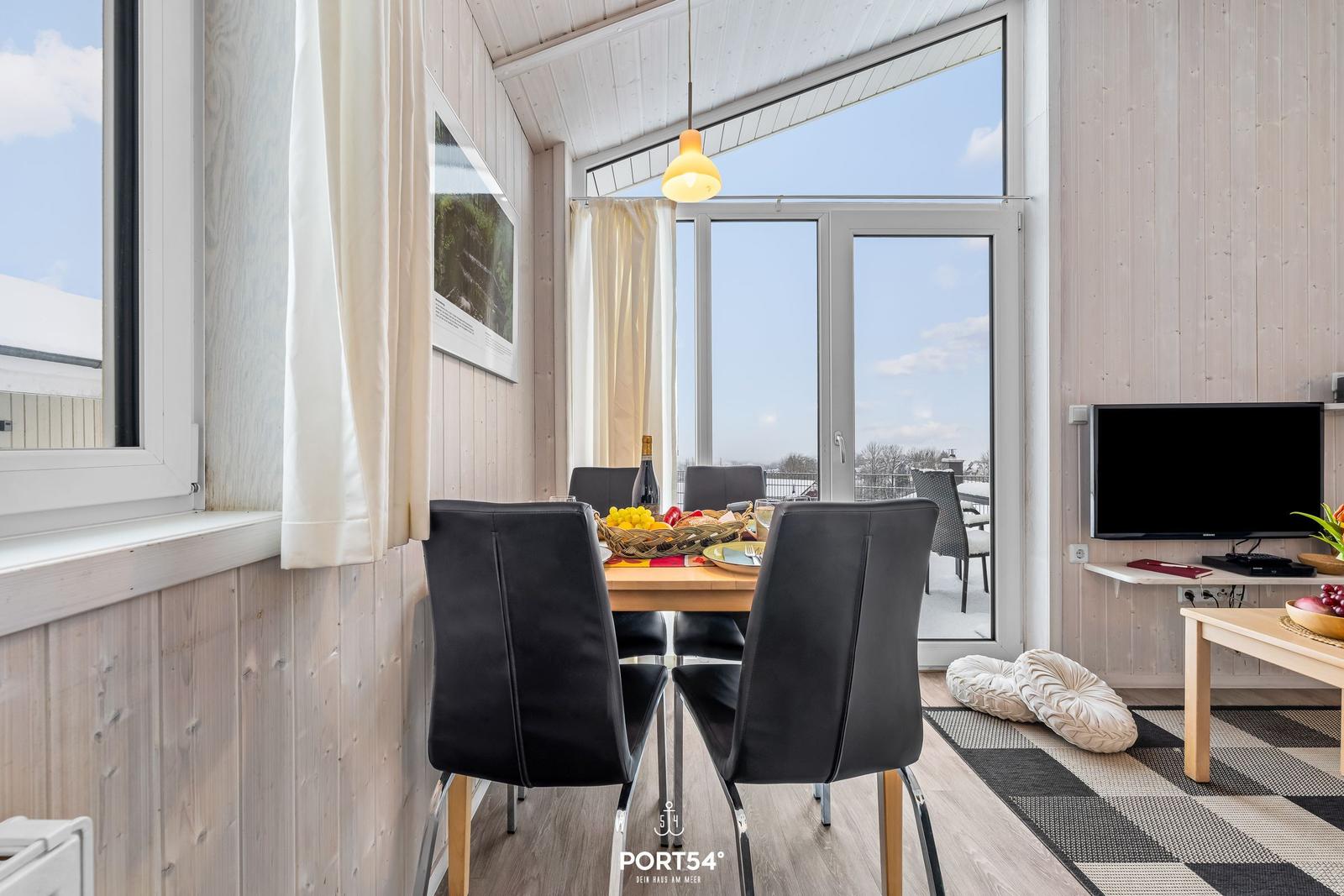 Essbereich mit Tisch, Stühlen und Blick auf Balkon. Holzwände und TV sichtbar.