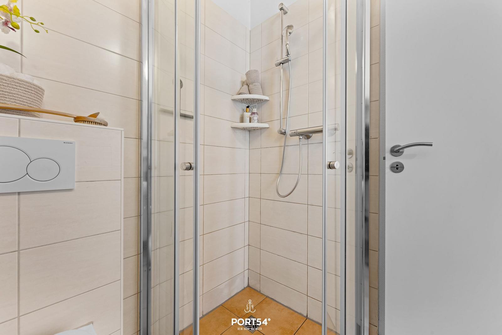 Duschkabine mit Glaswand, Duschbrause und Ablagefläche. WC und Badewanne sind ebenfalls vorhanden.