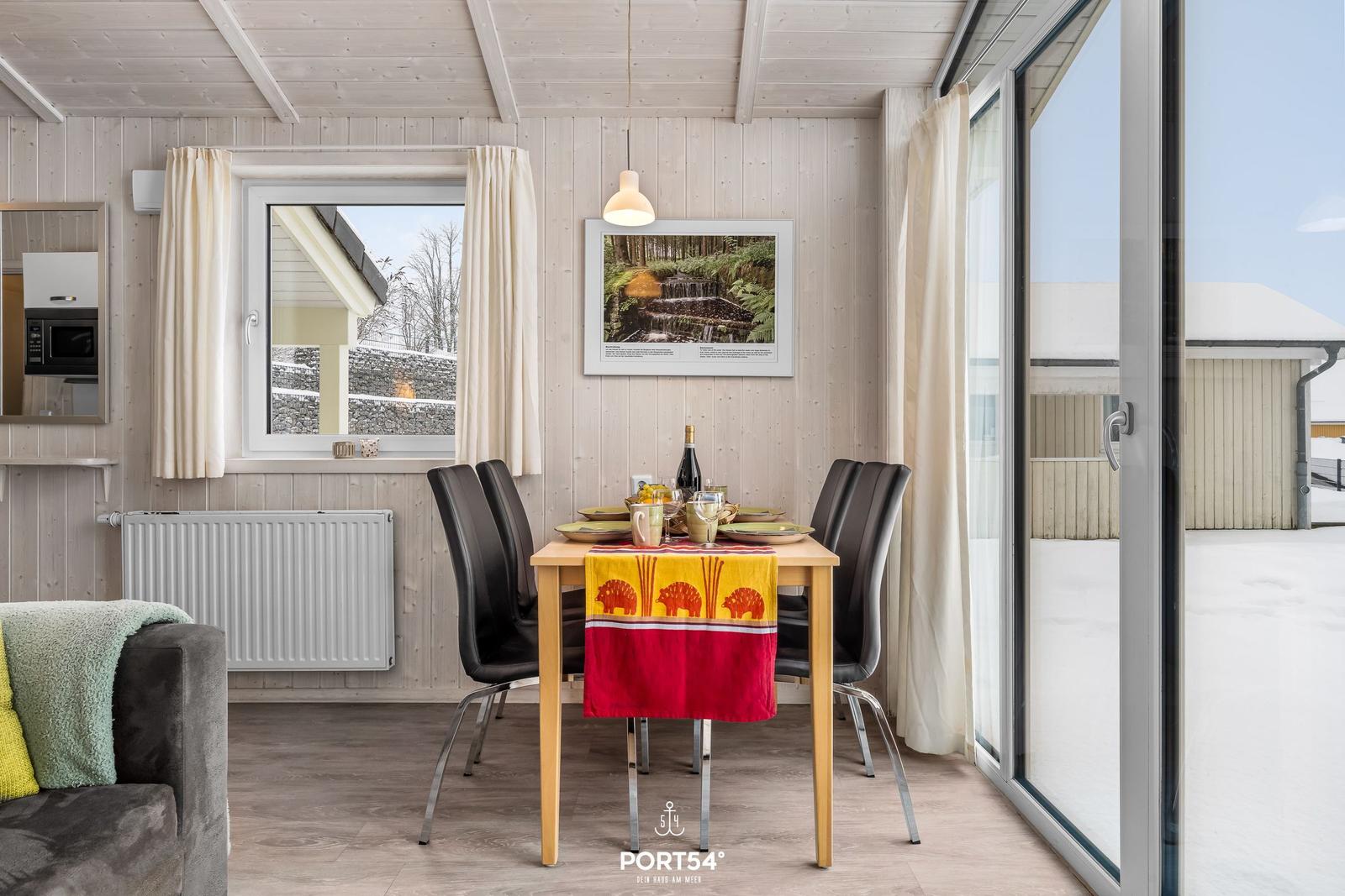 Esszimmer mit Tisch, Stühlen und Blick auf Schnee. Holzwand, Fenster und Glastür.