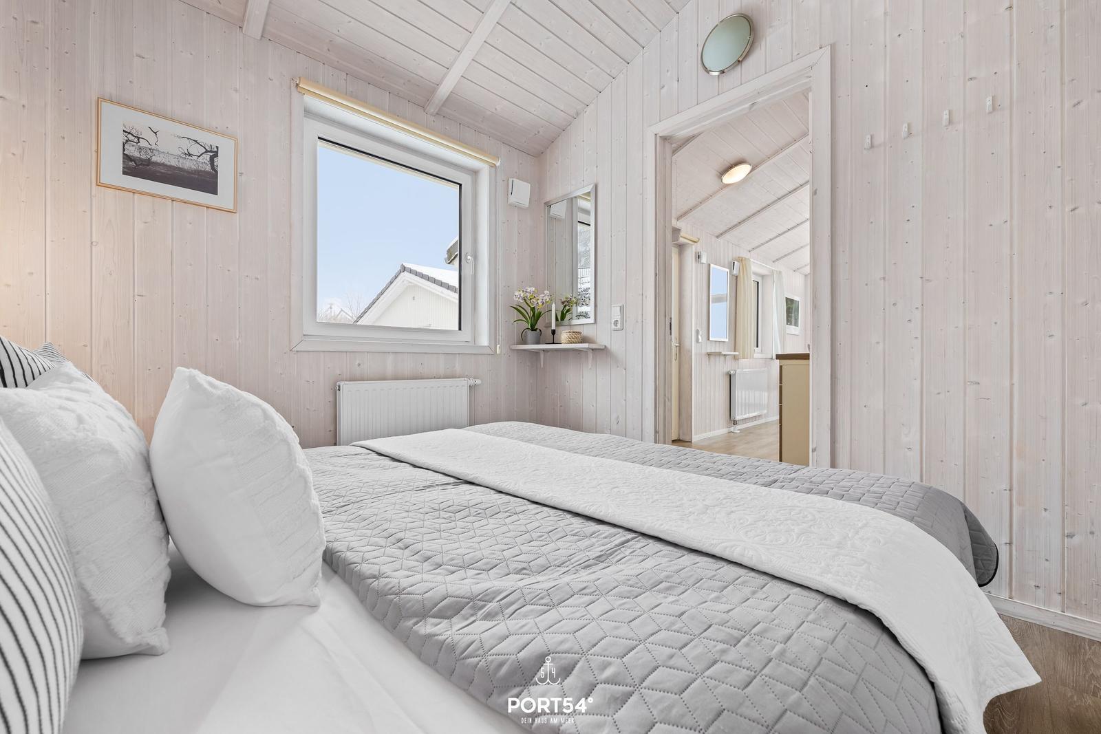Schlafzimmer mit Bett, Fenster und Holzwandverkleidung. Blick in angrenzenden Raum.