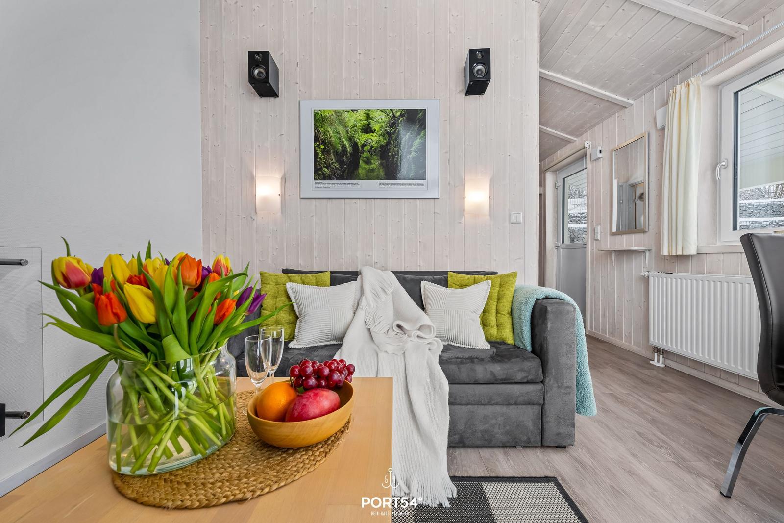 Wohnzimmer mit Sofa, Blumen und Obst auf Tisch. Holzwand und Fenster sichtbar.