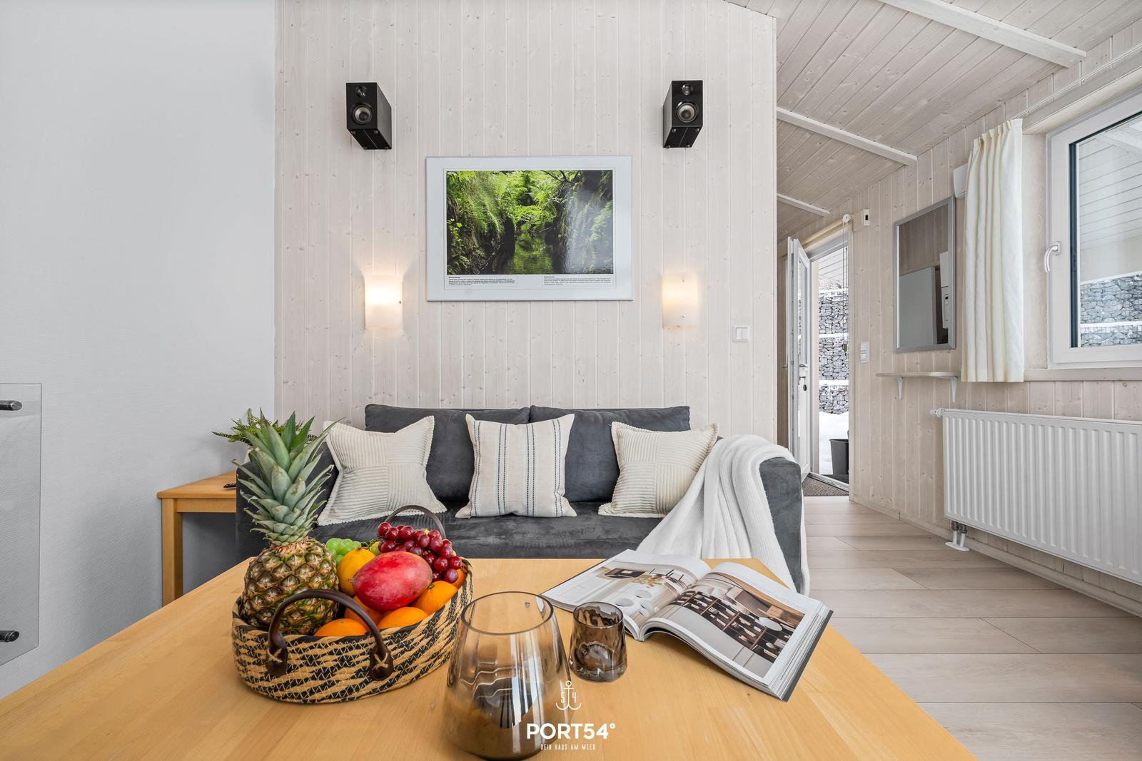 Living room with sofa, table, and fruit. Wall art and speakers are visible.
