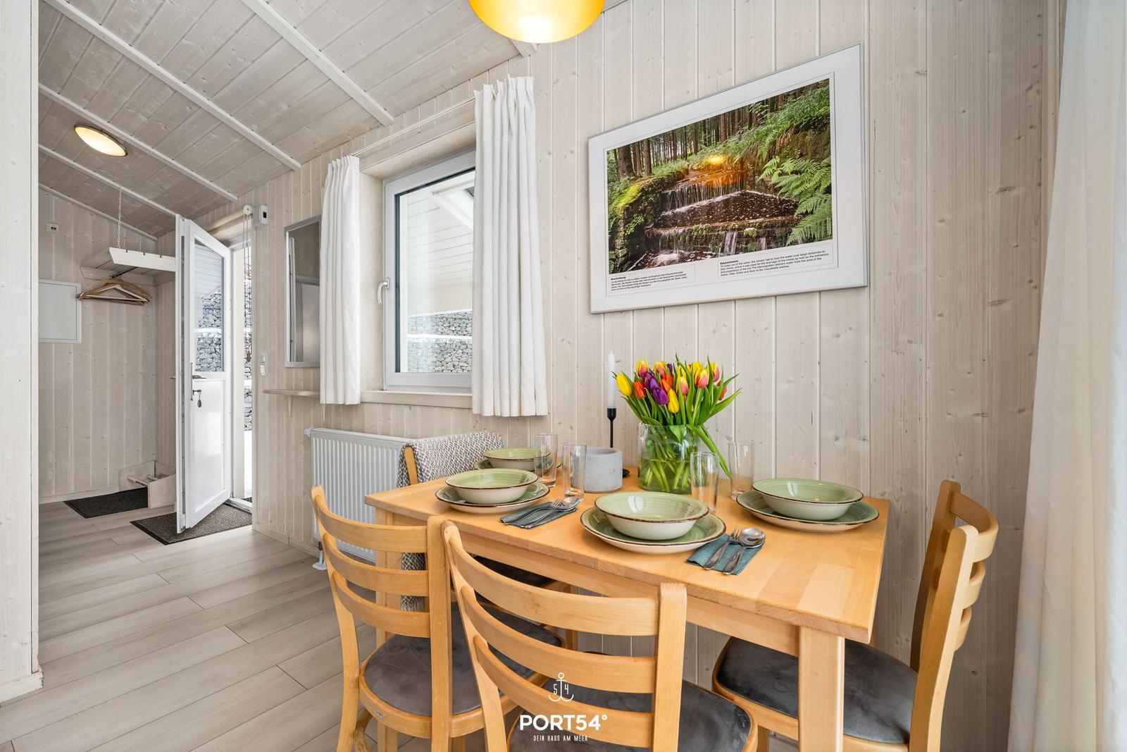 Dining room with wooden table, chairs, and flowers. Wall picture and window with curtains.