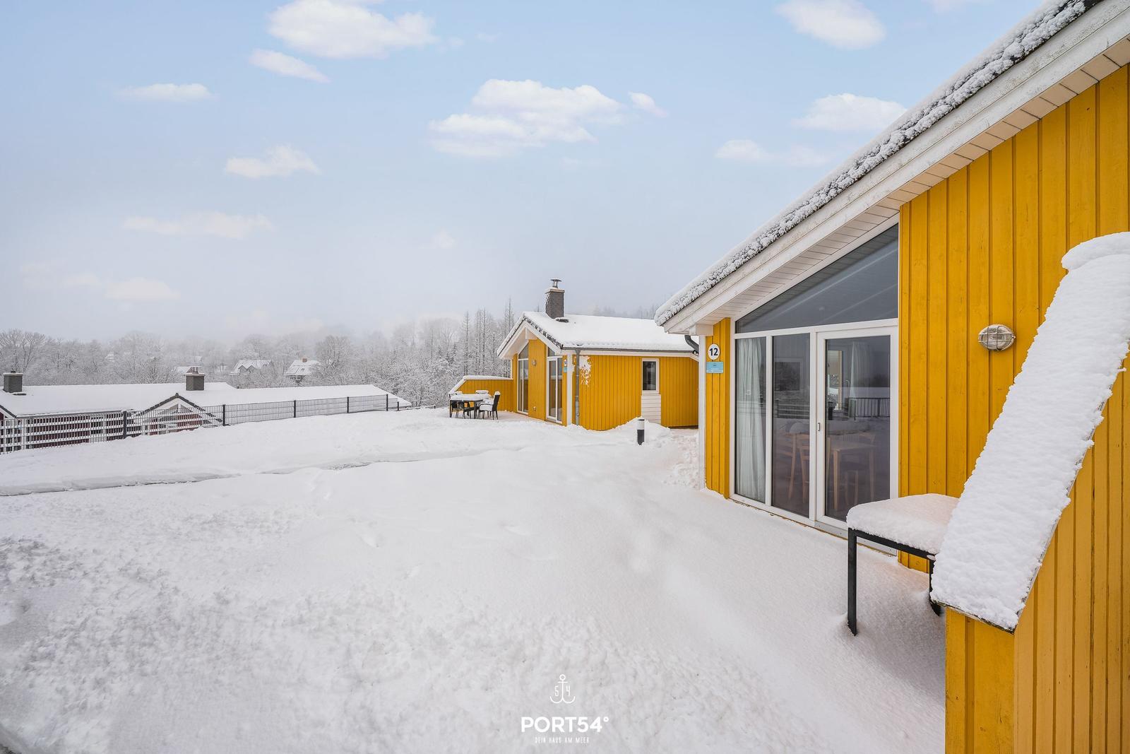 Snow covers the yard and yellow houses. A terrace with table and chairs is visible.