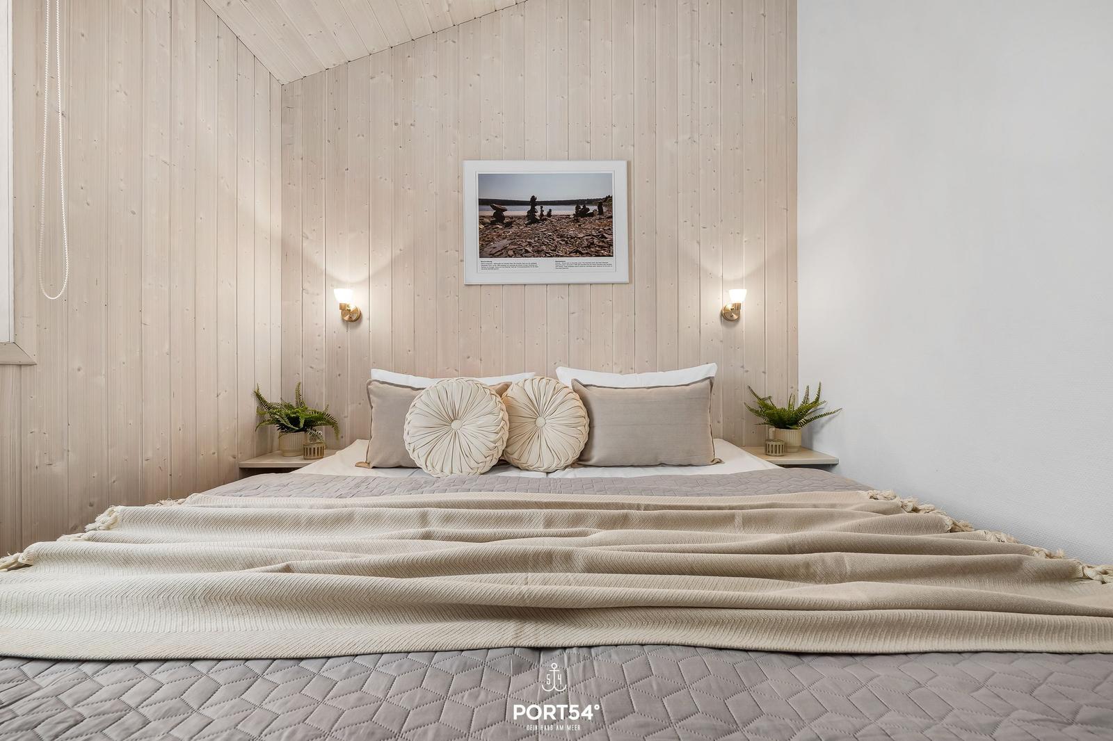 Bedroom with wooden wall, bed with pillows and blanket, wall picture and nightstands with plants.