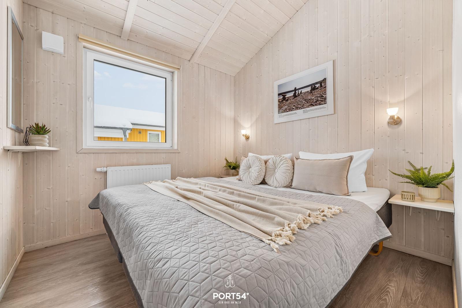 Bedroom with wooden walls, bed, and window. On the bed are pillows and a blanket.