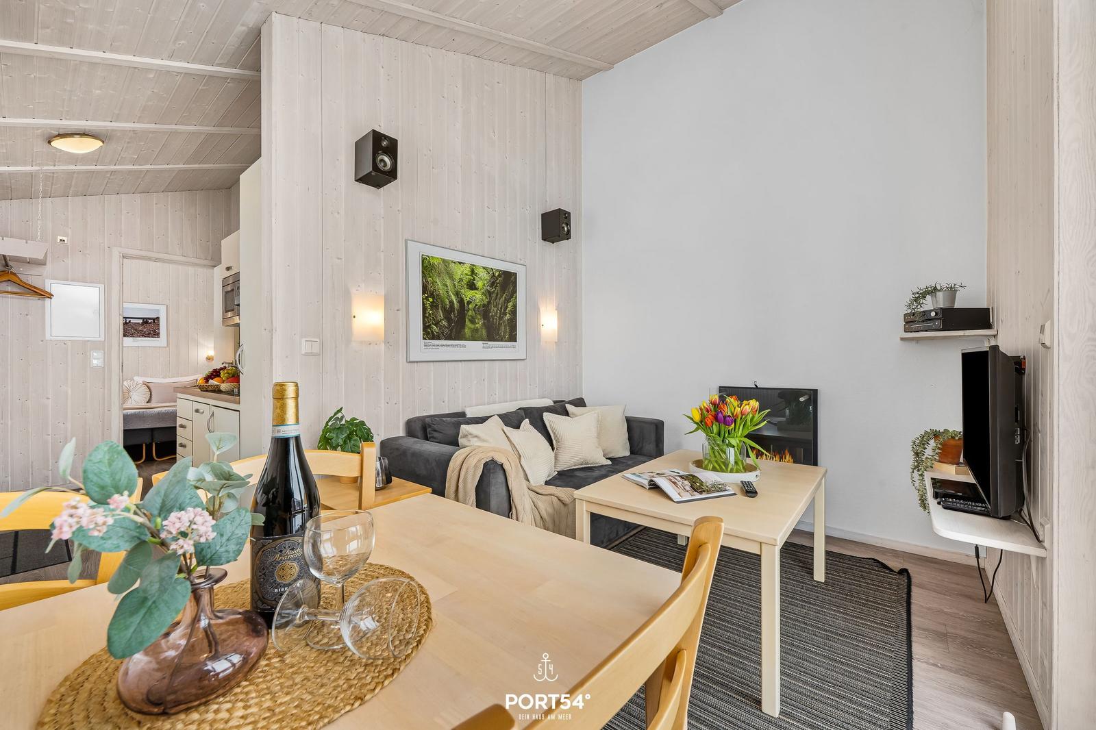 Living and dining area with sofa, table, and TV. Wooden walls and built-in kitchen visible.