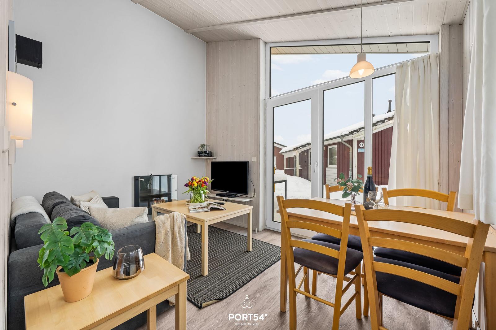 Living and dining area with sofa, table, chairs, and TV. Large window view to outdoor area.