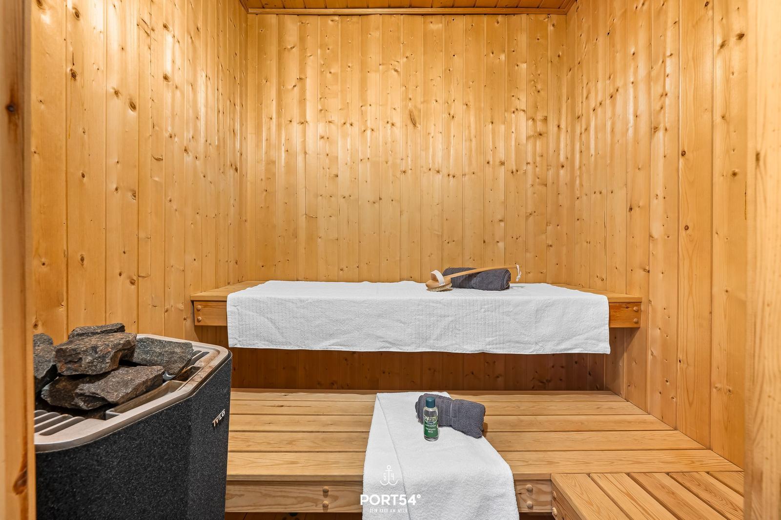 Sauna mit Holzwand, Liegefläche und Steinofen. Handtücher und Bürsten bereit.