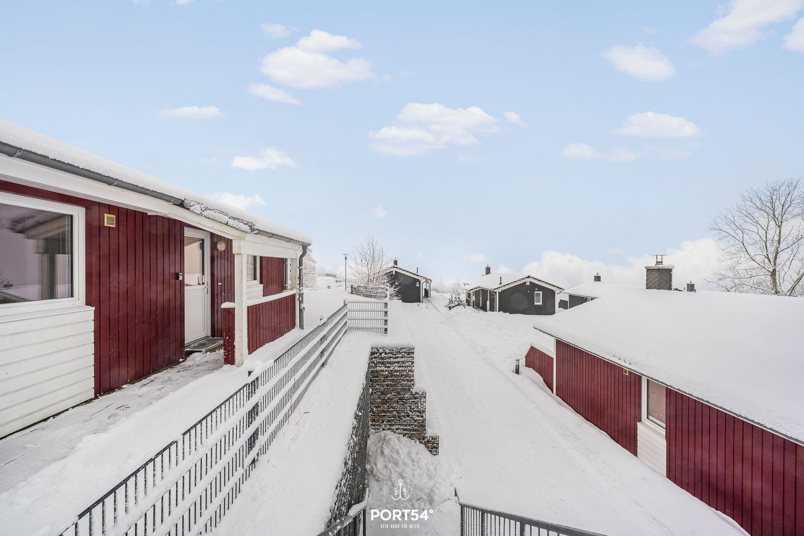 Schneebedeckte Holzhäuser mit roten Wänden und weißen Dächern in einer ruhigen Winterlandschaft.