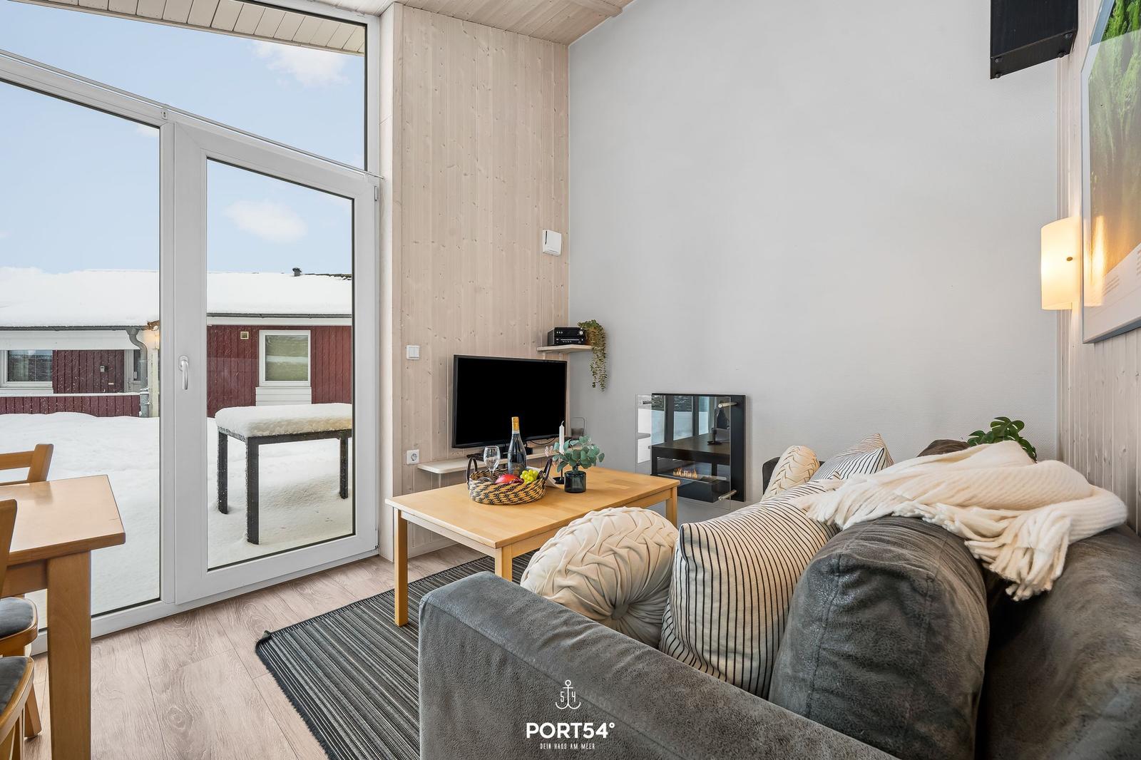 Wohnzimmer mit Sofa, Fernseher und Kamin. Durchgehende Glasfront zum verschneiten Außenbereich.