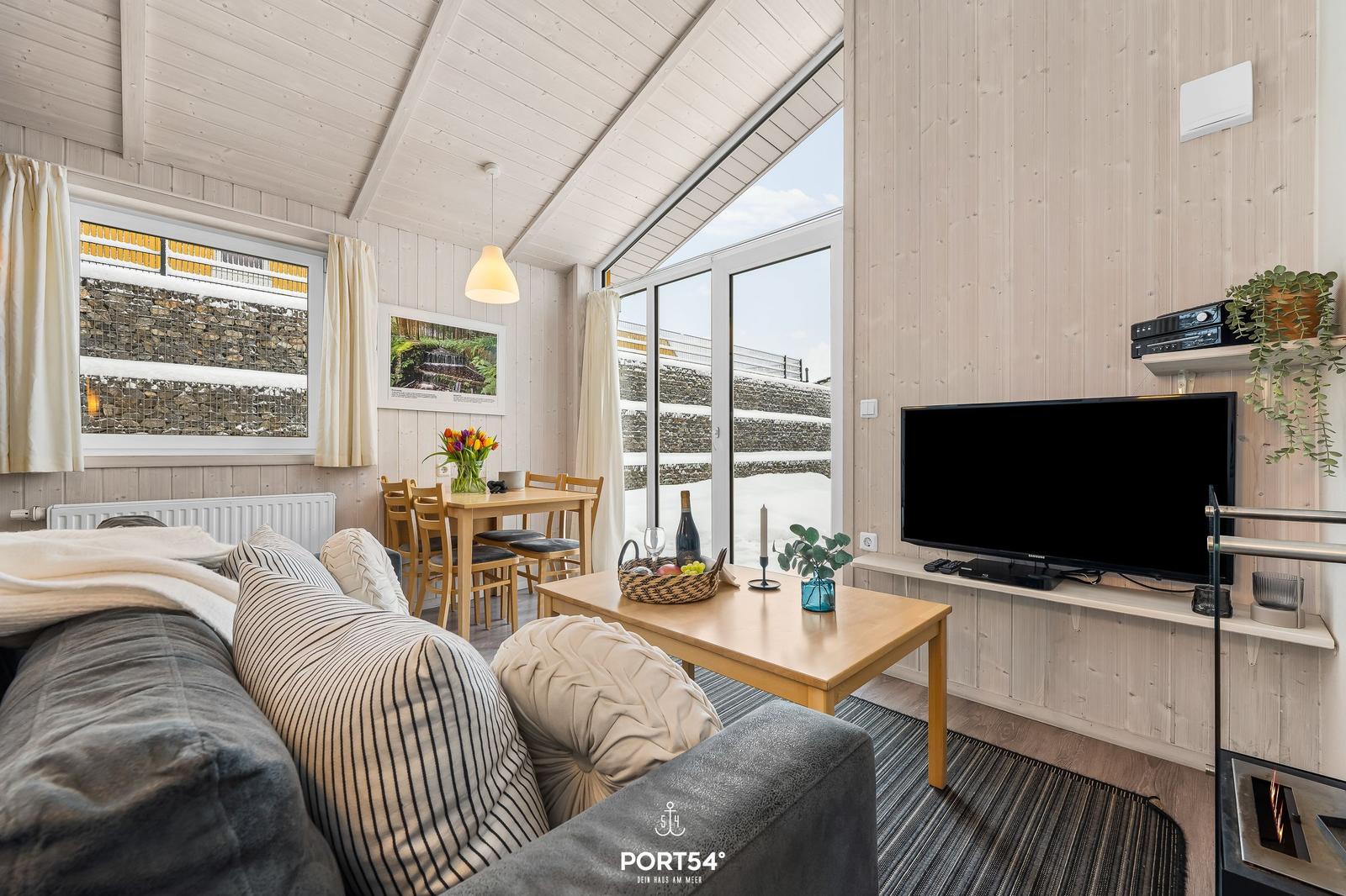 Wohnzimmer mit Sofa, Esstisch und Fernseher. Große Fenster mit Blick auf Steinmauer.