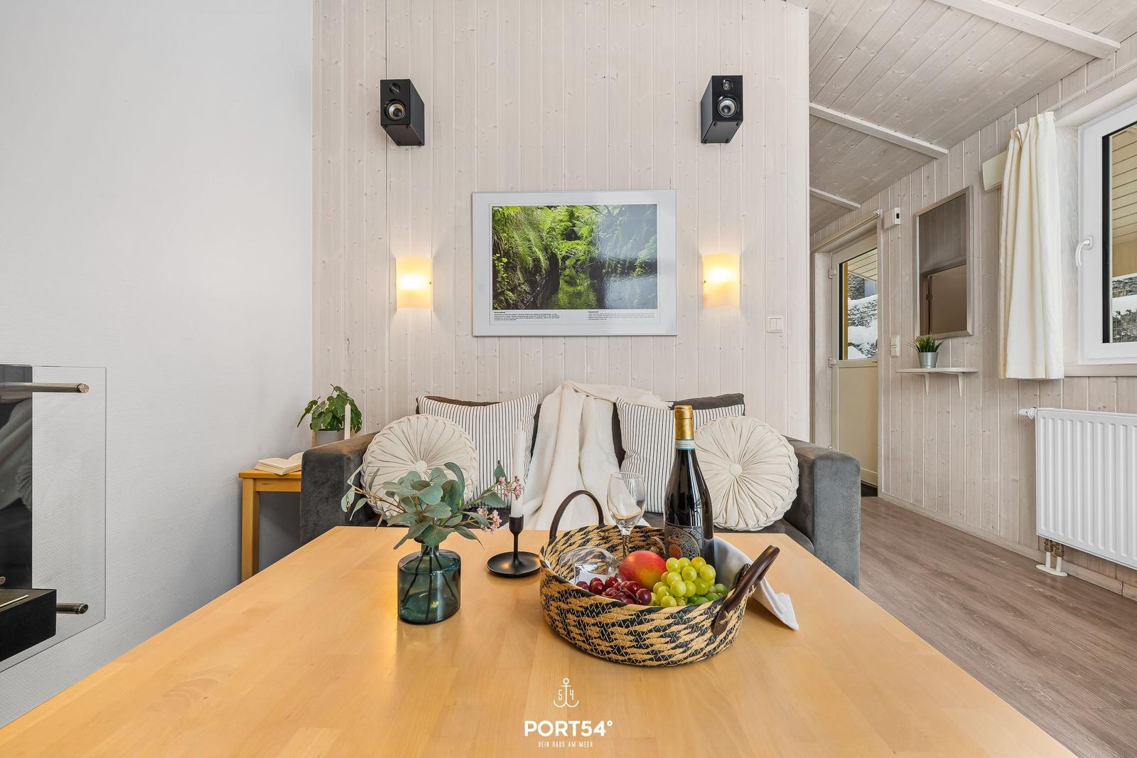 Esstisch mit Obstkorb, Sofa und Wandbild in einem Raum mit Holzwänden.
