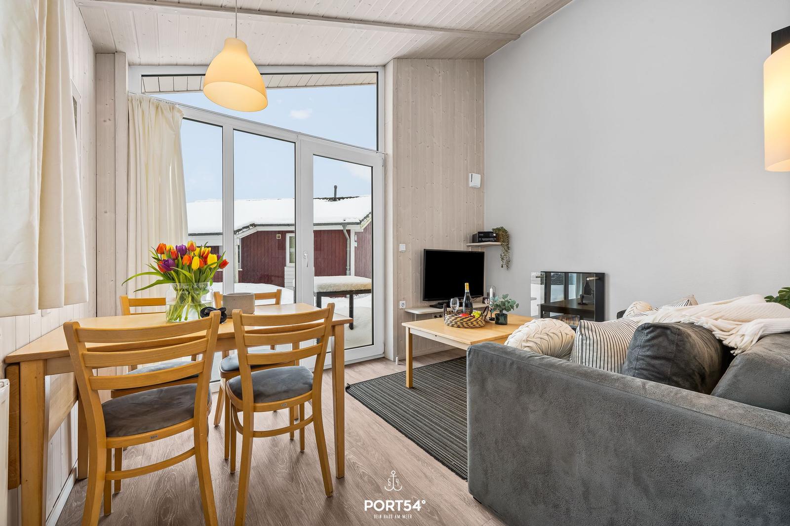 Wohn- und Essbereich mit Sofa, Tisch und Stühlen. Durchgang zur Terrasse mit Blick auf rote Gebäude.