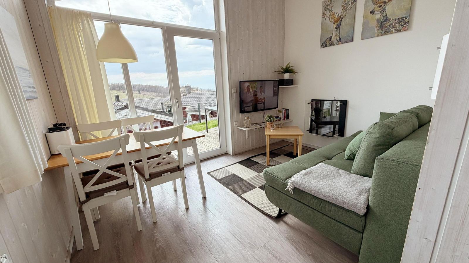 Wohn- und Essbereich mit grünem Sofa, Tisch mit Stühlen und Balkon mit Blick.