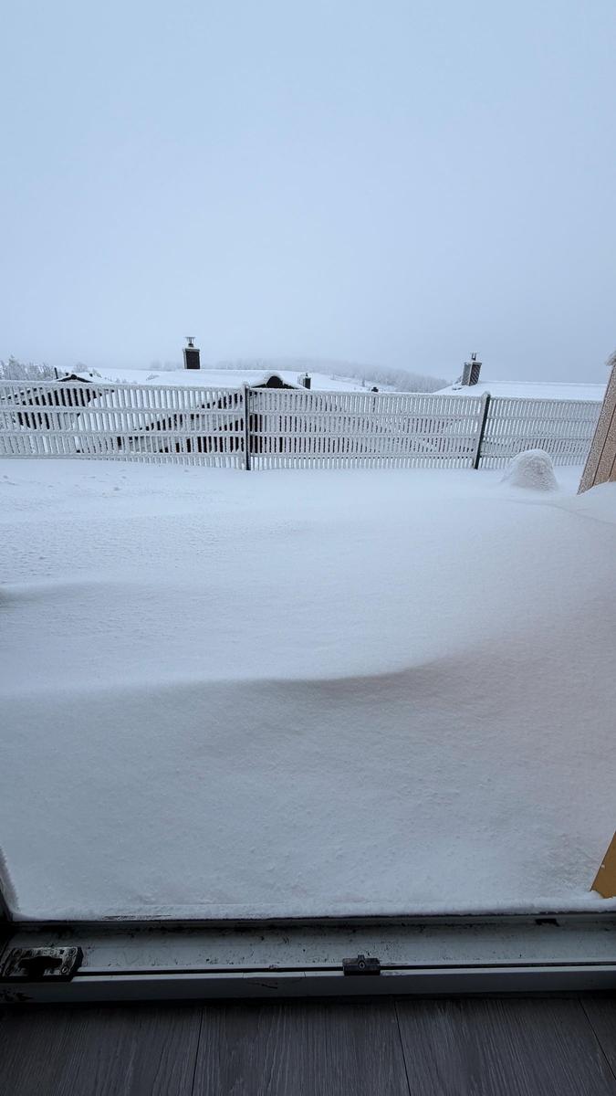 Schneebedeckter Balkon mit weißem Zaun und verschneiten Gebäuden im Hintergrund.