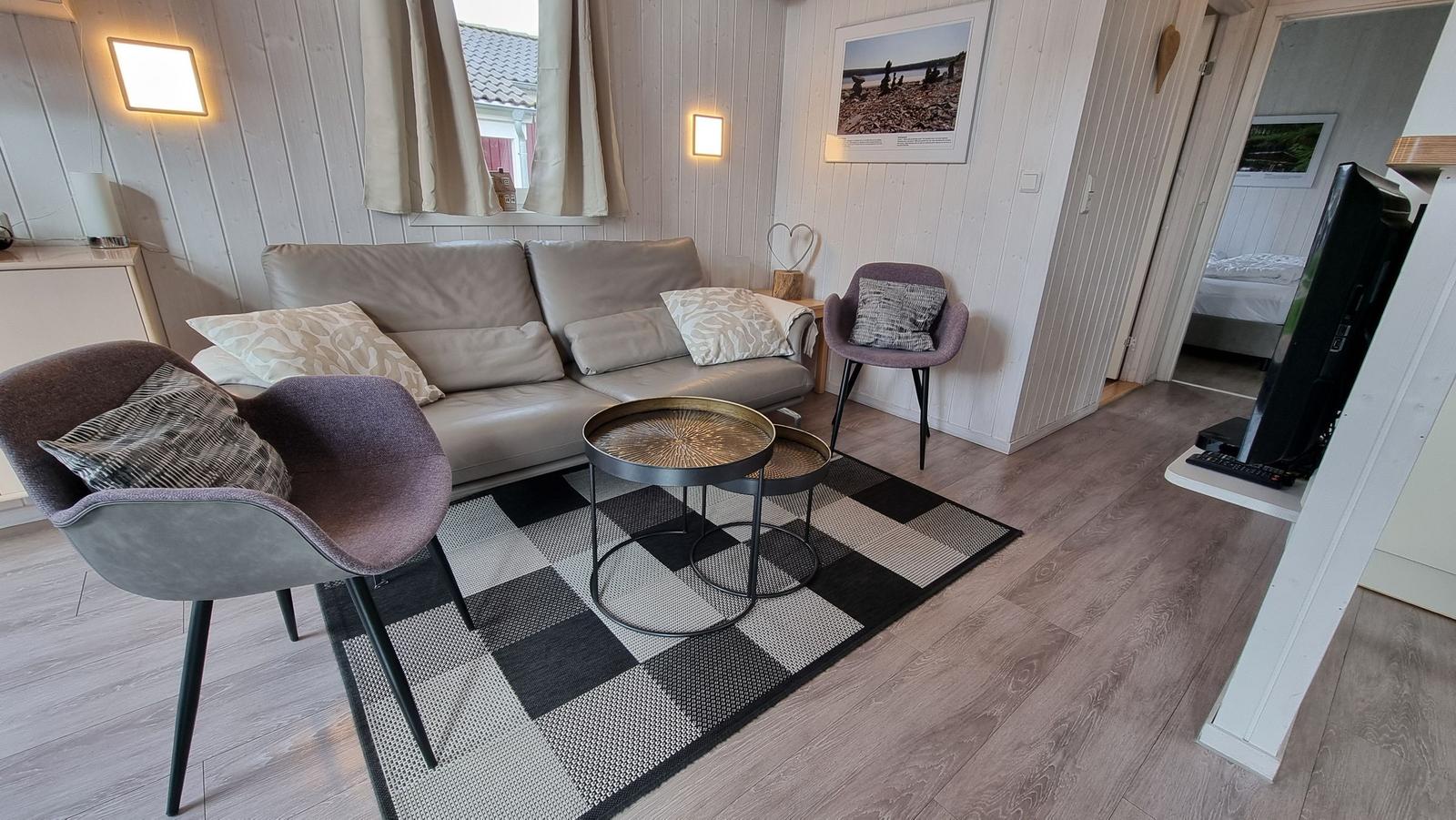Wohnzimmer mit Sofa, zwei Sesseln, Tisch und Teppich. Durchgang zur Schlafzimmer.