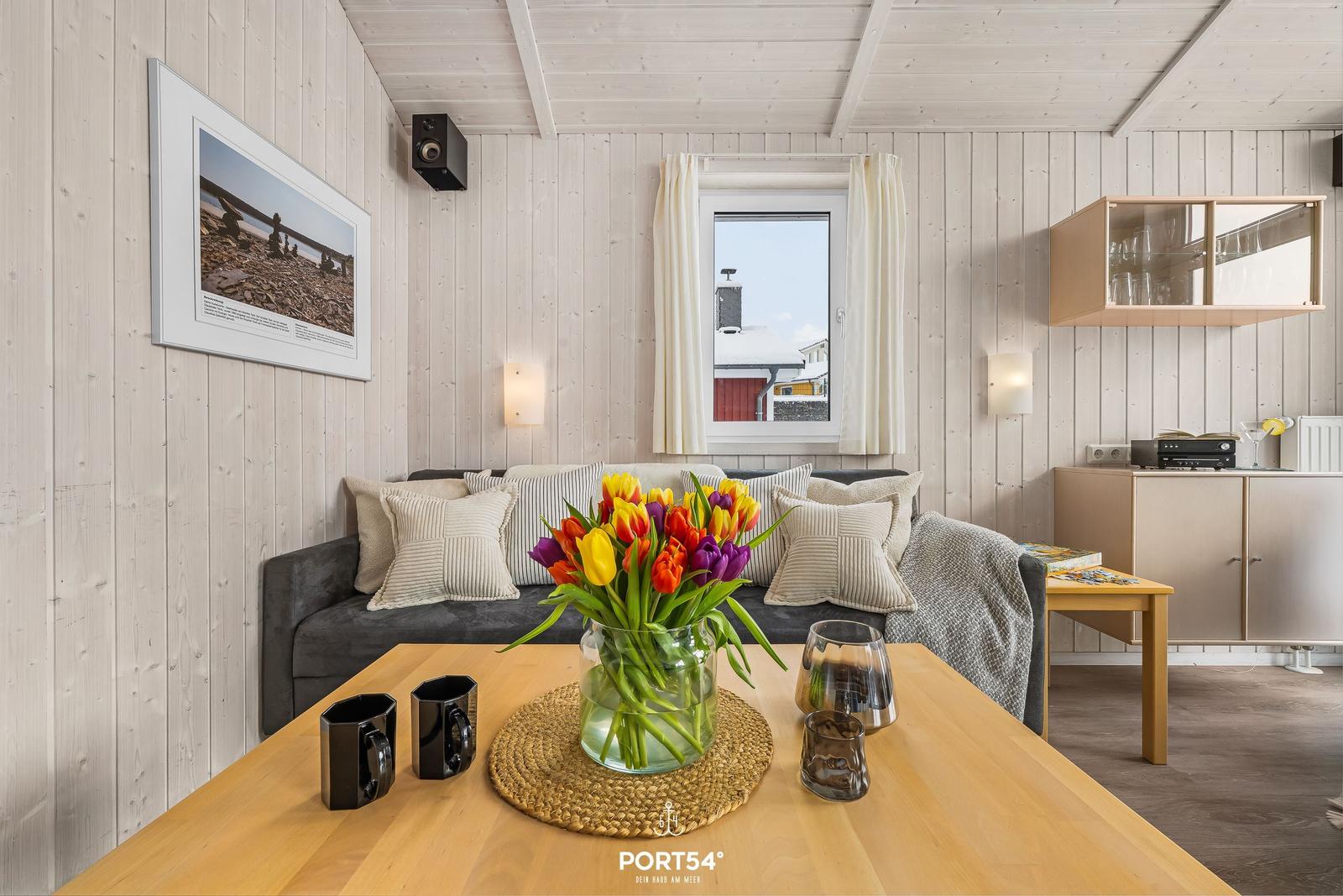 Wohnzimmer mit Sofa, Tisch und Blumen. Fenster zeigt Außenansicht.