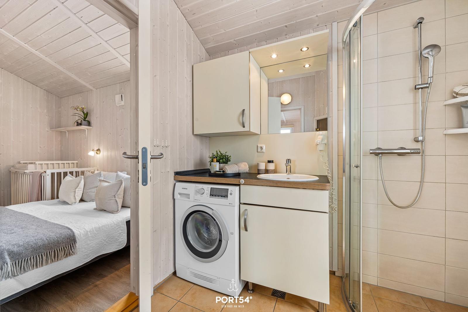 Badezimmer mit Dusche, Waschmaschine und Spiegel. Schlafzimmer mit Bett und Babybett sichtbar.