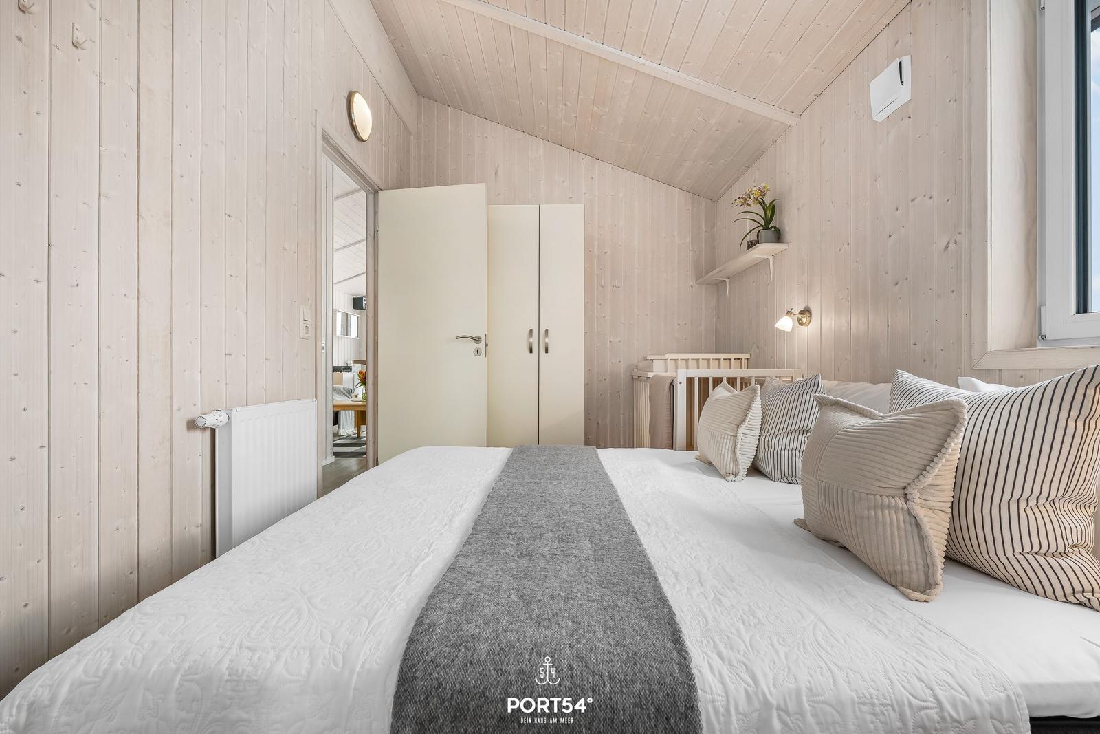 Schlafzimmer mit Bett, Kleiderschrank und Babybett. Holzwände und Fenster sichtbar.