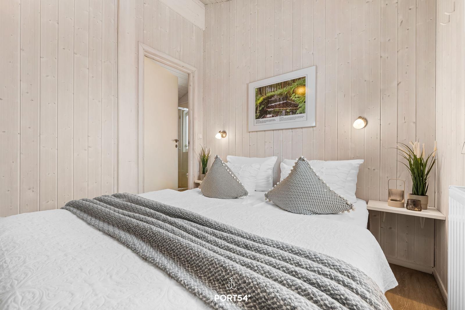Schlafzimmer mit Bett, Holzwand und Wandbild. Neben dem Bett steht ein kleiner Tisch mit Pflanze.