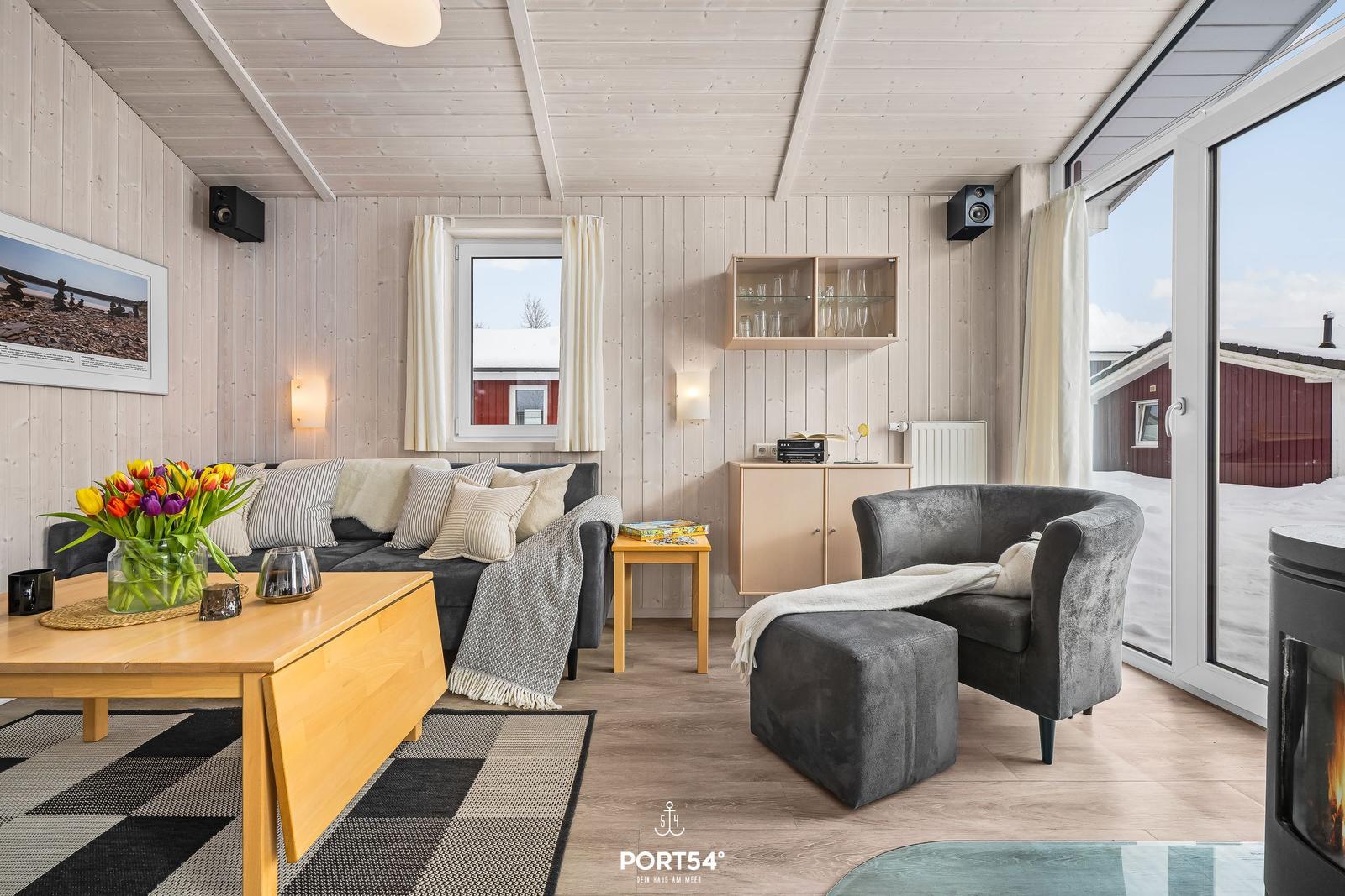 Wohnzimmer mit Sofa, Sessel und Kamin. Durchgehende Glasfront mit Blick auf Schnee.