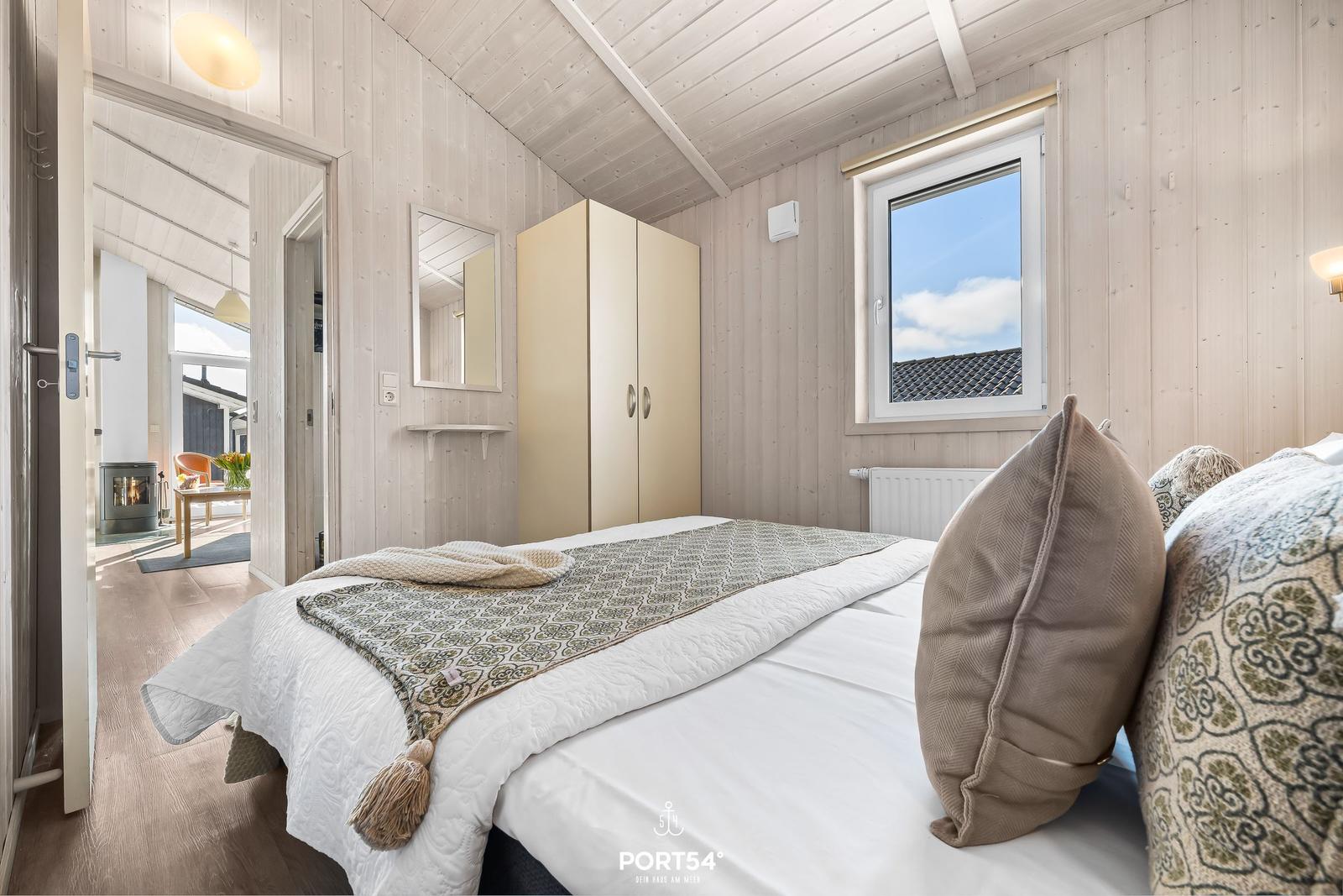 Schlafzimmer mit Bett, Kleiderschrank und Fenster. Durchgang zu Wohnbereich mit Kamin.