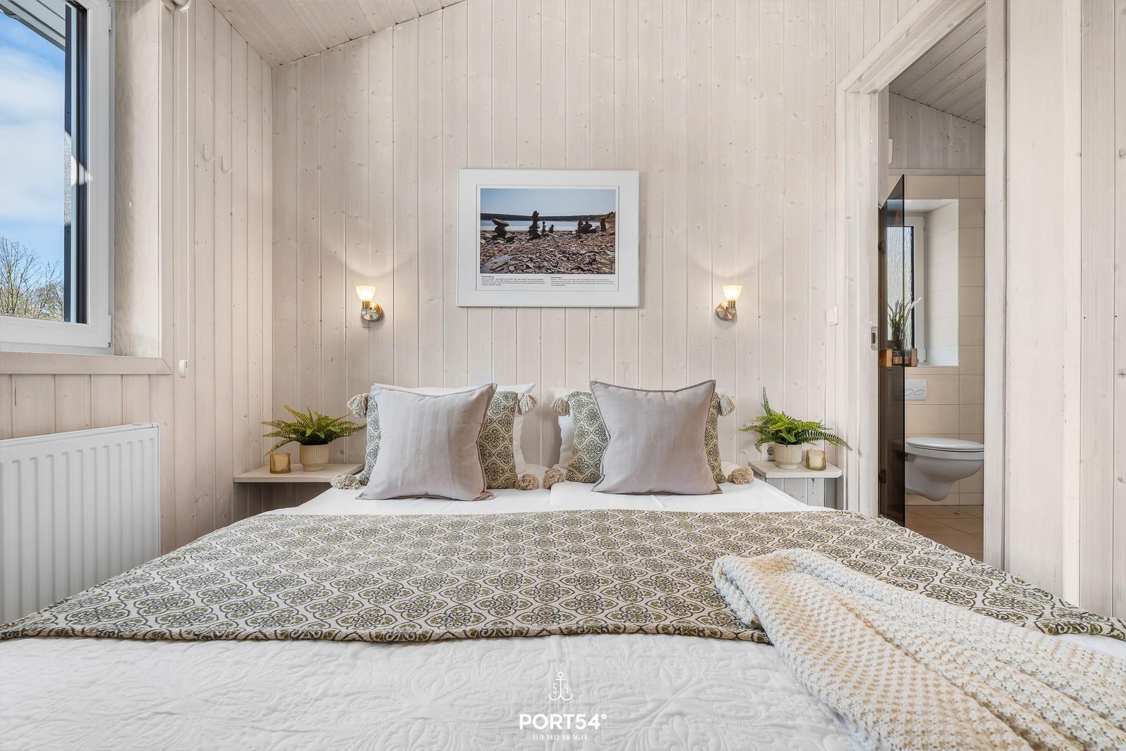 Schlafzimmer mit Bett, Wandbild und Pflanzen. Badezimmer im Hintergrund sichtbar.
