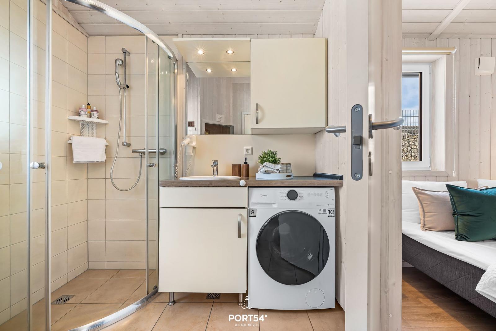 Badezimmer mit Dusche, Waschmaschine und Spiegel. Blick in Schlafzimmer mit Bett.
