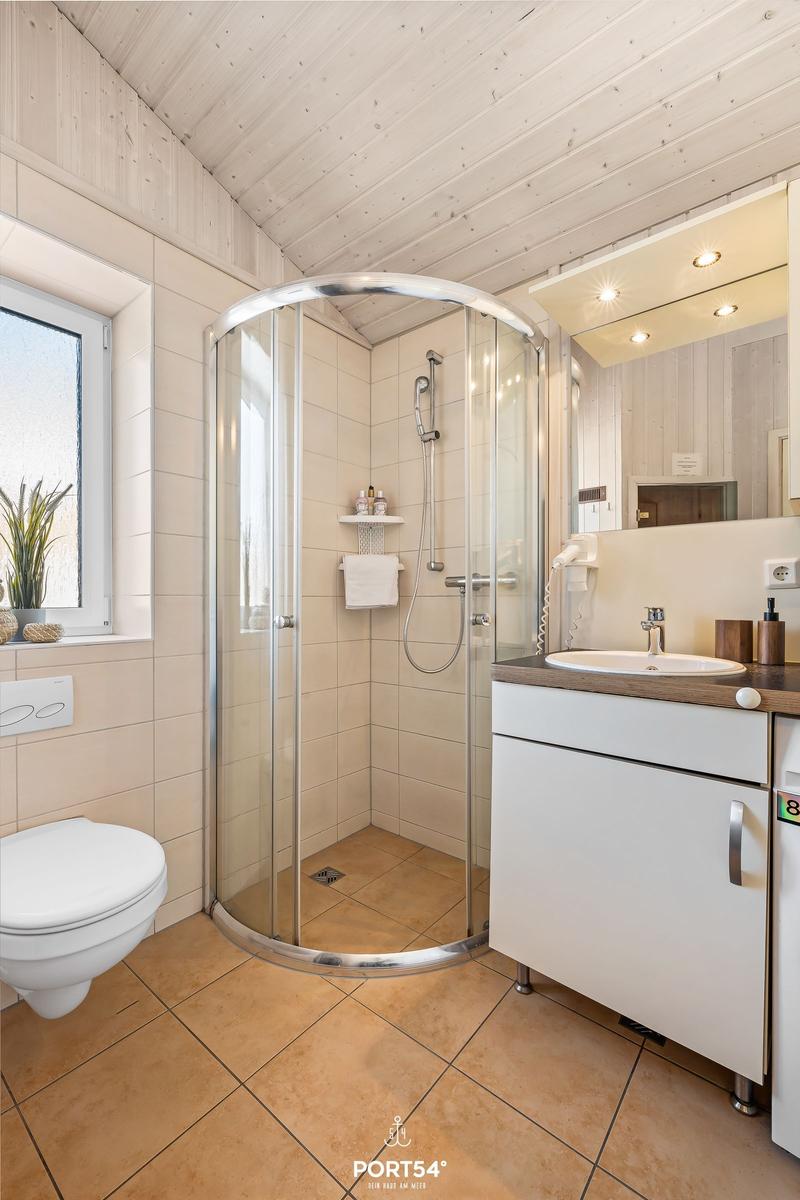 Badezimmer mit Dusche, Waschbecken und Toilette. Holzdecke und Fliesenboden.
