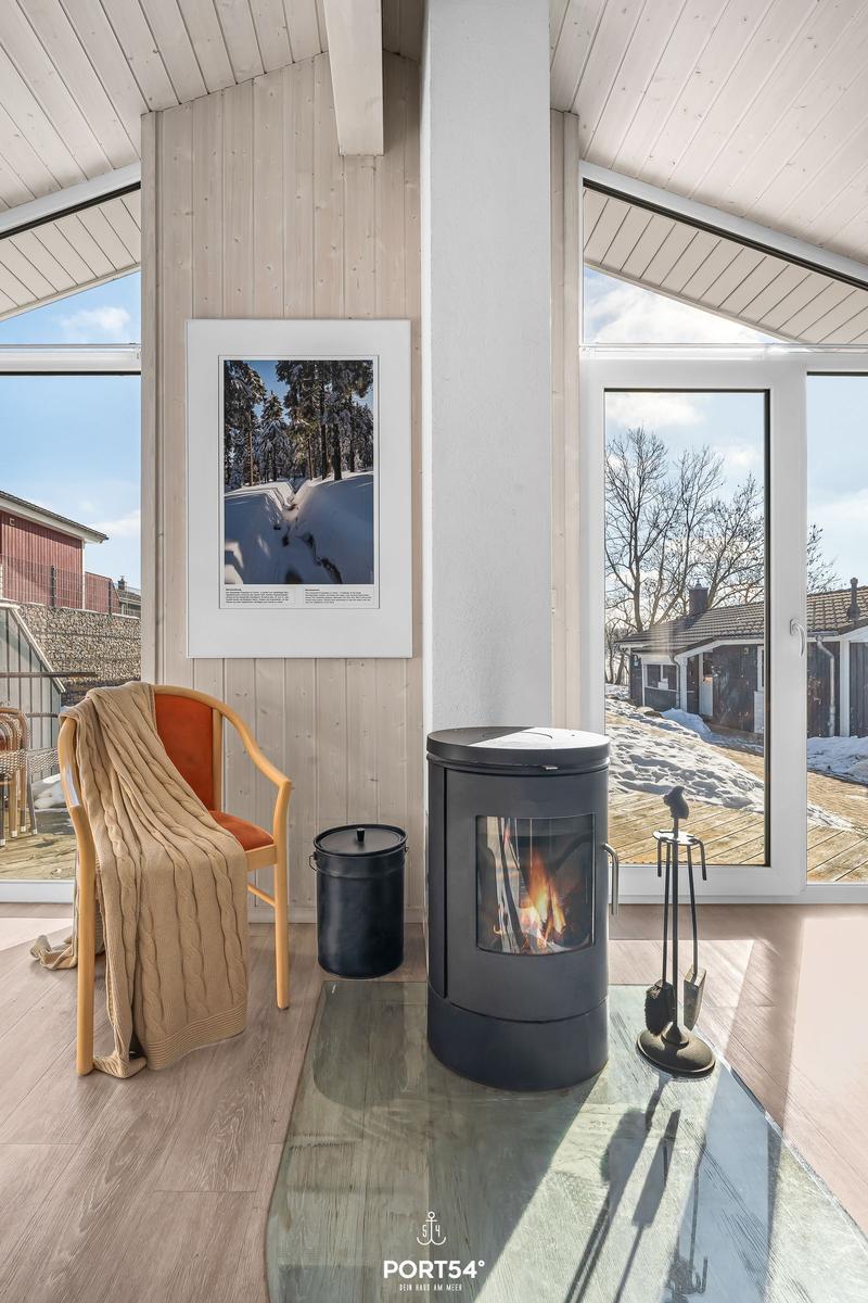 Raum mit Kamin, Sessel und Glasplatte. Große Fenster zeigen Außenbereich.