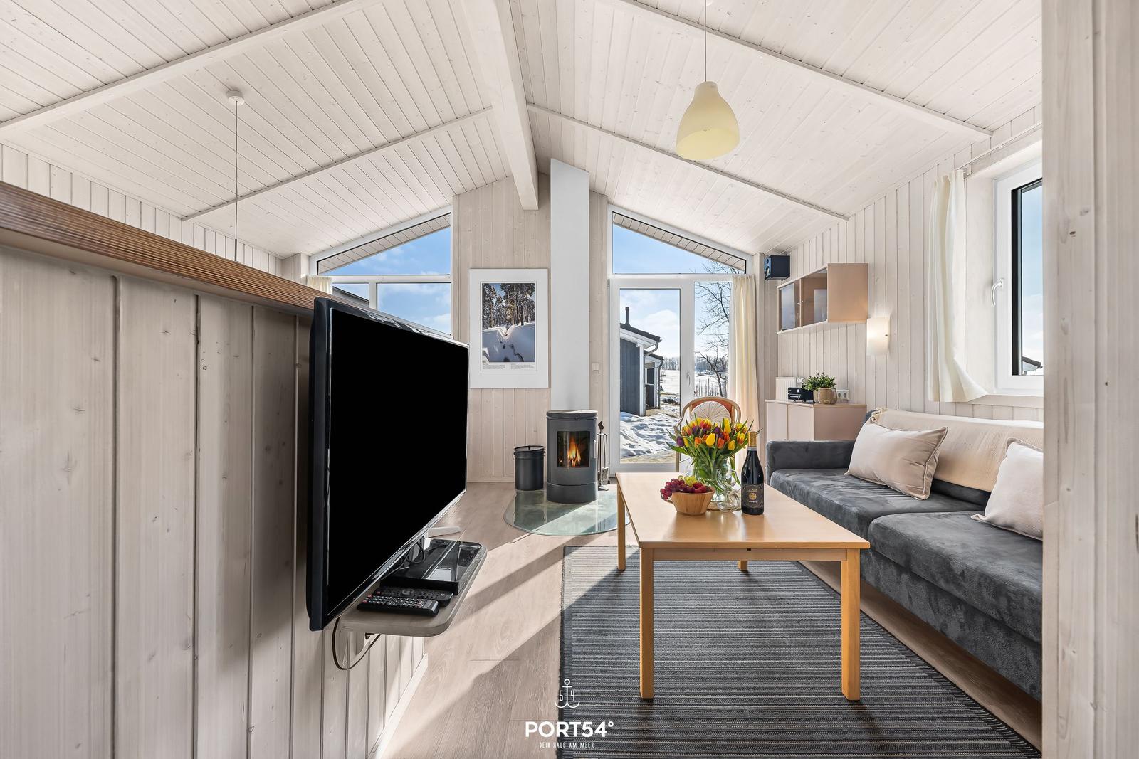 Wohnzimmer mit Sofa, Couchtisch und Fernseher. Holzwände und große Fenster.