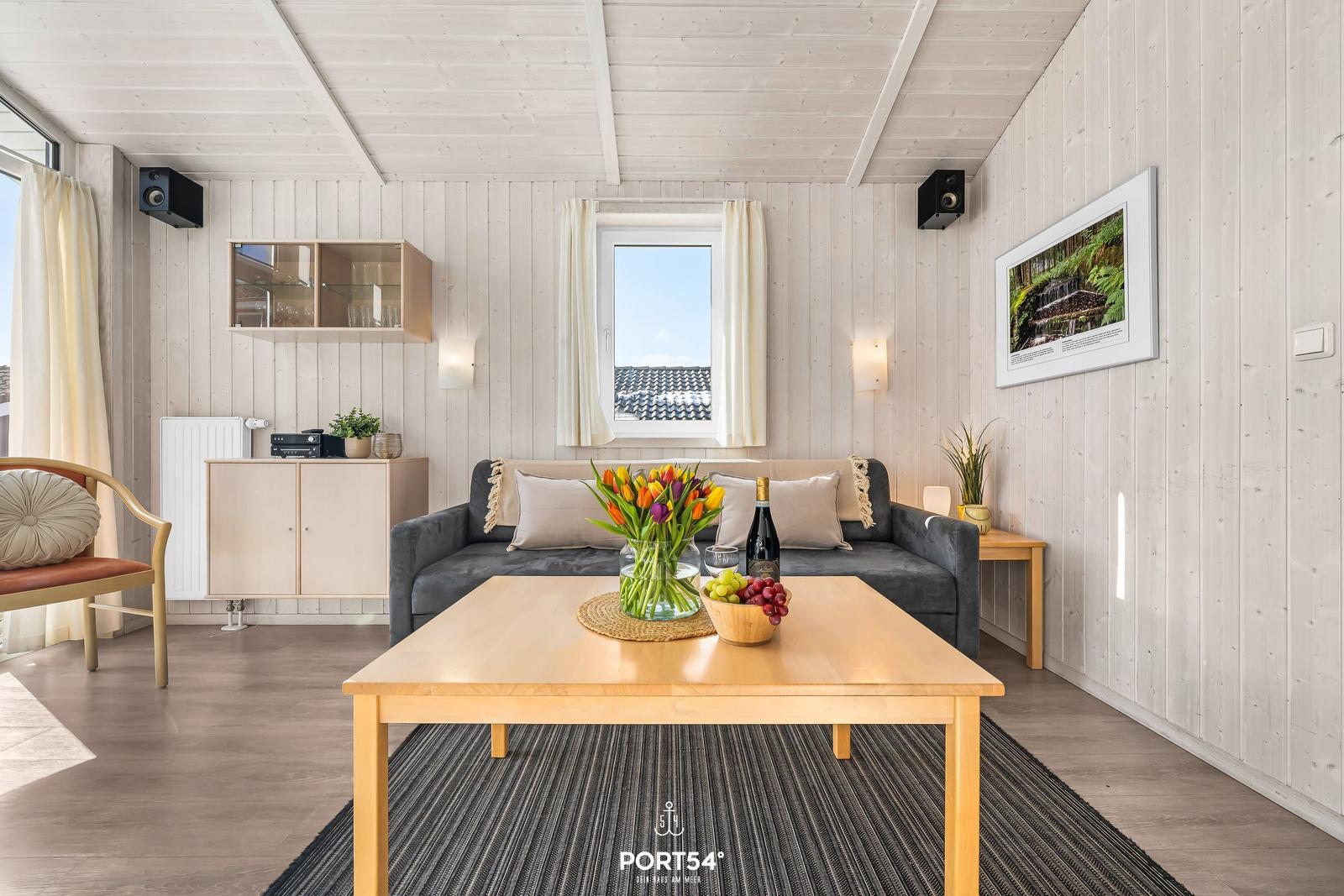 Wohnzimmer mit Sofa, Tisch und Fenster. Holzwände und -decke. Blumen und Obst auf dem Tisch.