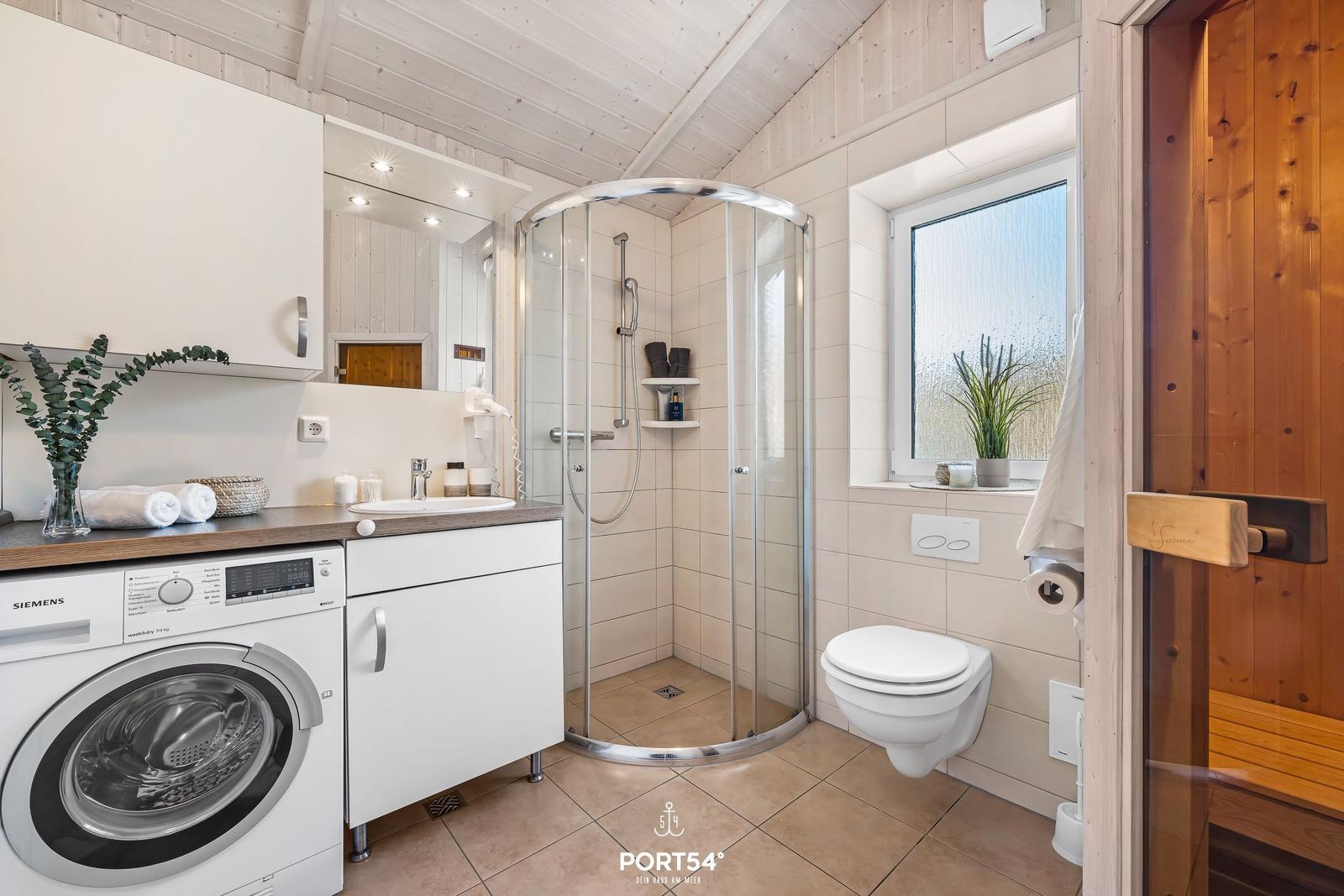 Badezimmer mit Dusche, Waschmaschine und Toilette. Fenster mit Pflanze.