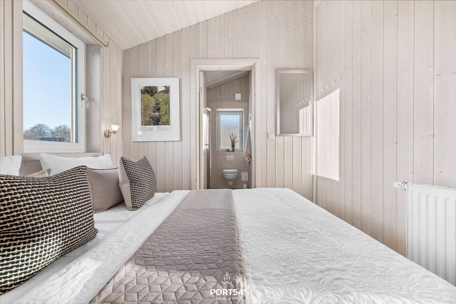 Schlafzimmer mit Holzwand, Bett, Fenster und angrenzender Toilette.