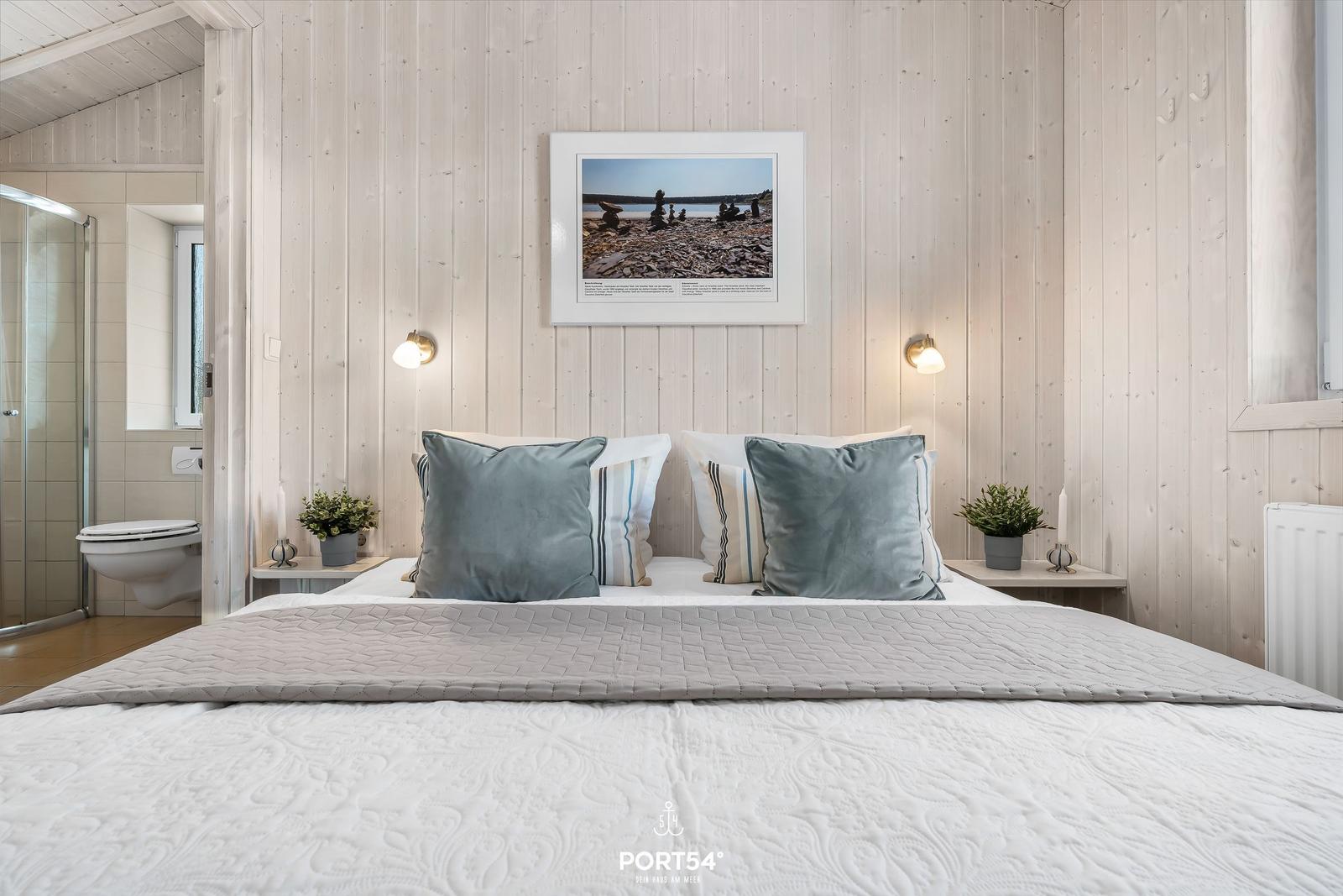 Schlafzimmer mit Bett, Wandbild und Nachttischen. Badezimmer im Hintergrund.