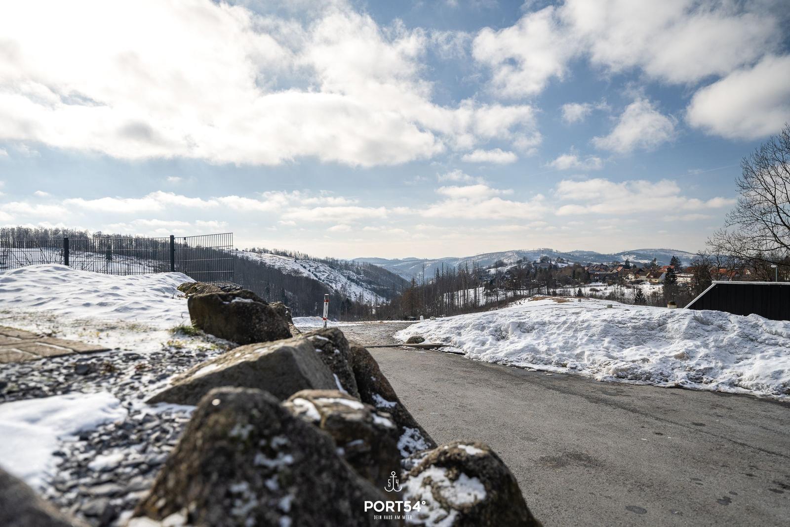 Schneebedeckter Weg mit Steinmauer und Blick auf bewaldete Hügel und Ferienhaus.