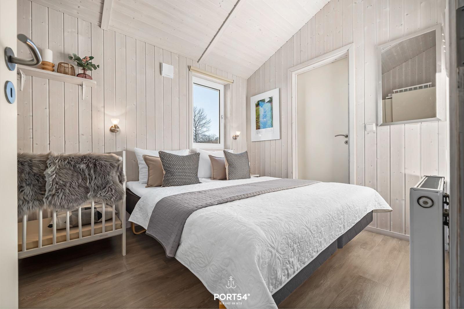 Schlafzimmer mit Bett, Fenster und Holzwandverkleidung. Links Sitzbank mit Felldecke.