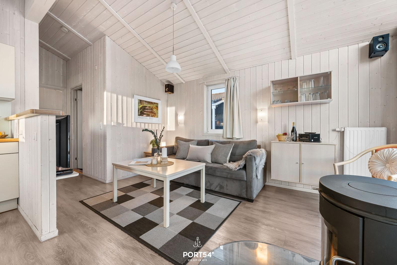 Wohnzimmer mit Sofa, Tisch und Kaminofen. Holzwände und -decke. Fenster mit Vorhang.