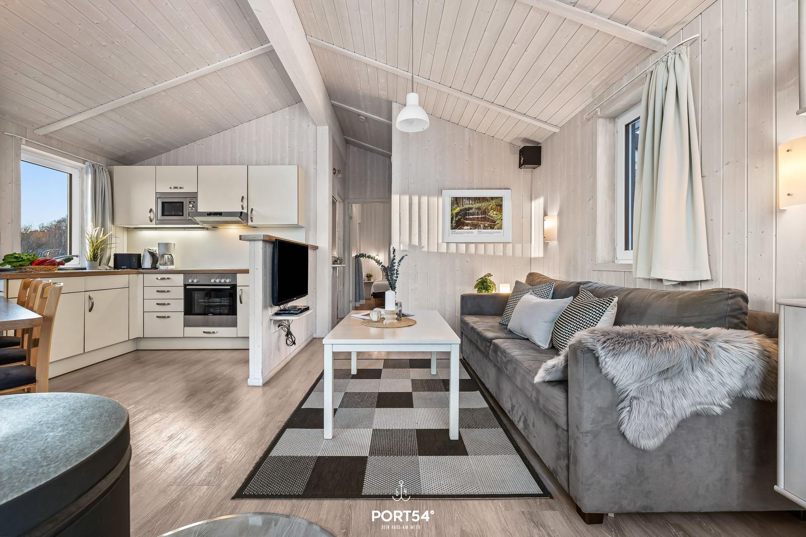 Wohnbereich mit Küche, Sofa und Tisch. Holzdecke und -wände. Fenster mit Vorhängen.