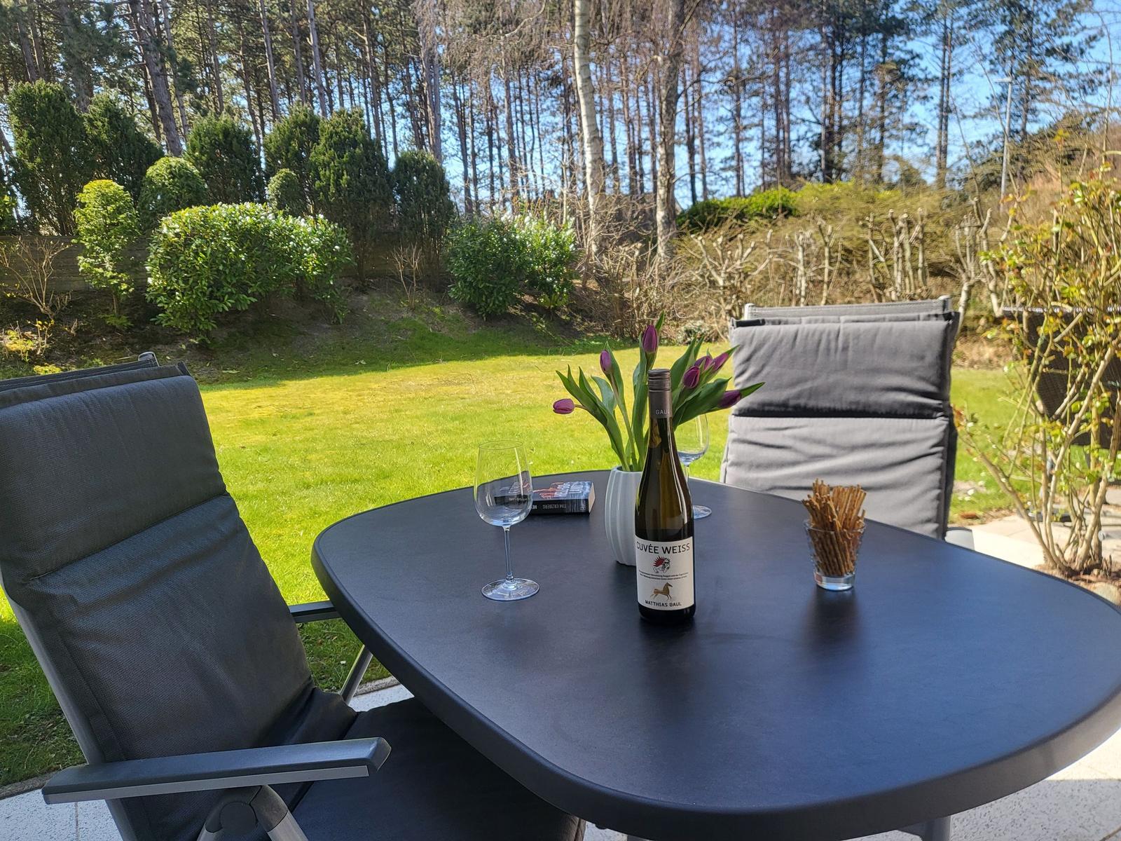 Außenbereich mit Tisch, Stühlen, Weinflasche und Tulpen. Grasfläche und Wald im Hintergrund.