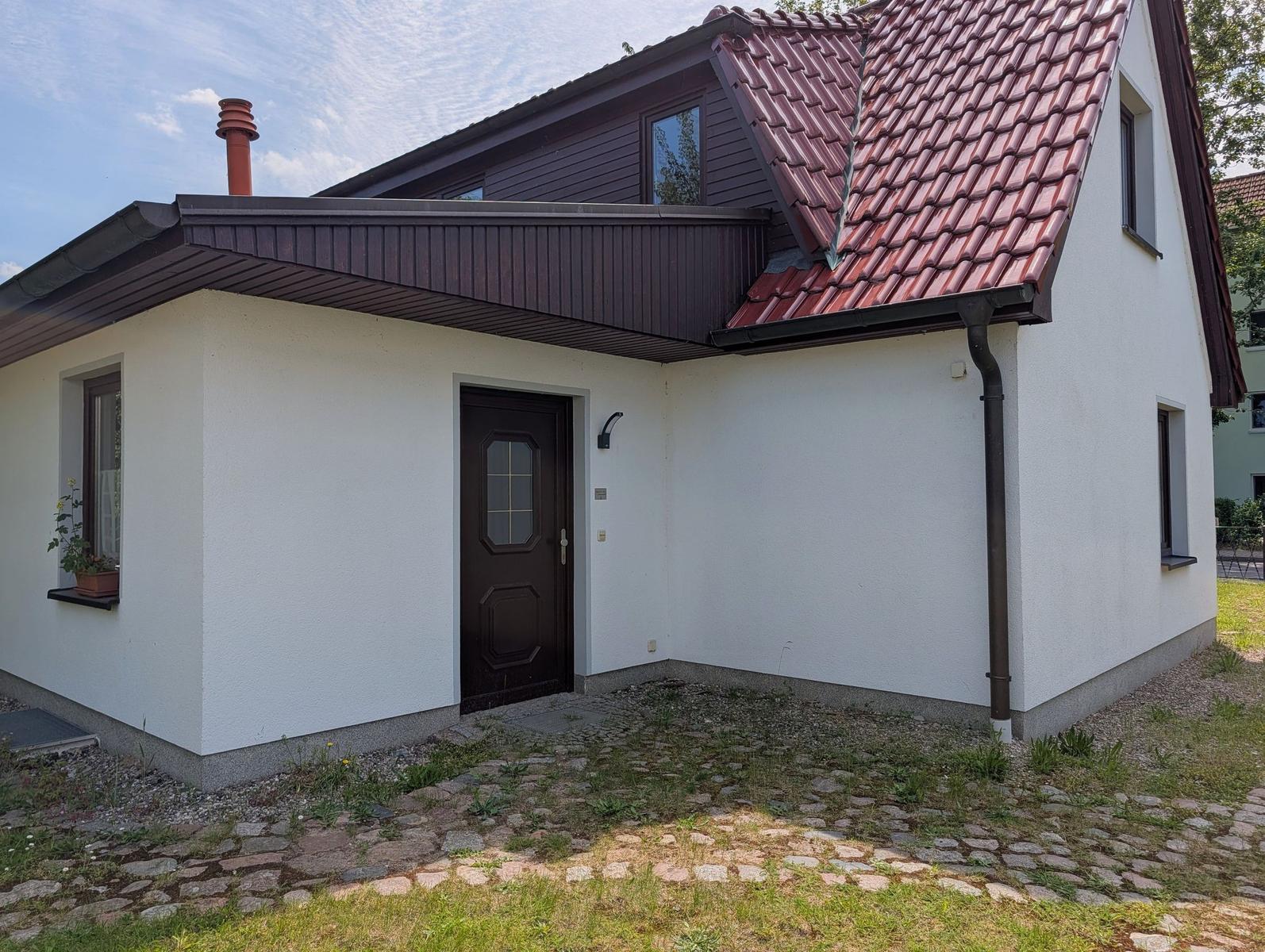 Haus mit weißer Fassade, braunem Dach und Eingangstür. Vor dem Haus befindet sich ein gepflasterter Bereich.